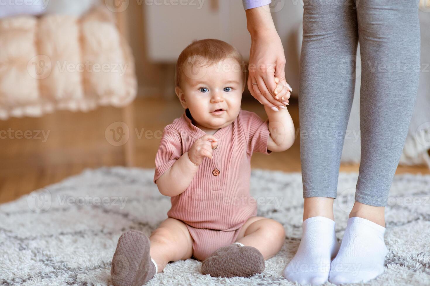 Adorable little kid holding mom hand, sitting on floor, got tired while making first steps, copy space. Beautiful blonde baby sitting on carpet at home, holding mother hand, smiling at camera, closeup photo