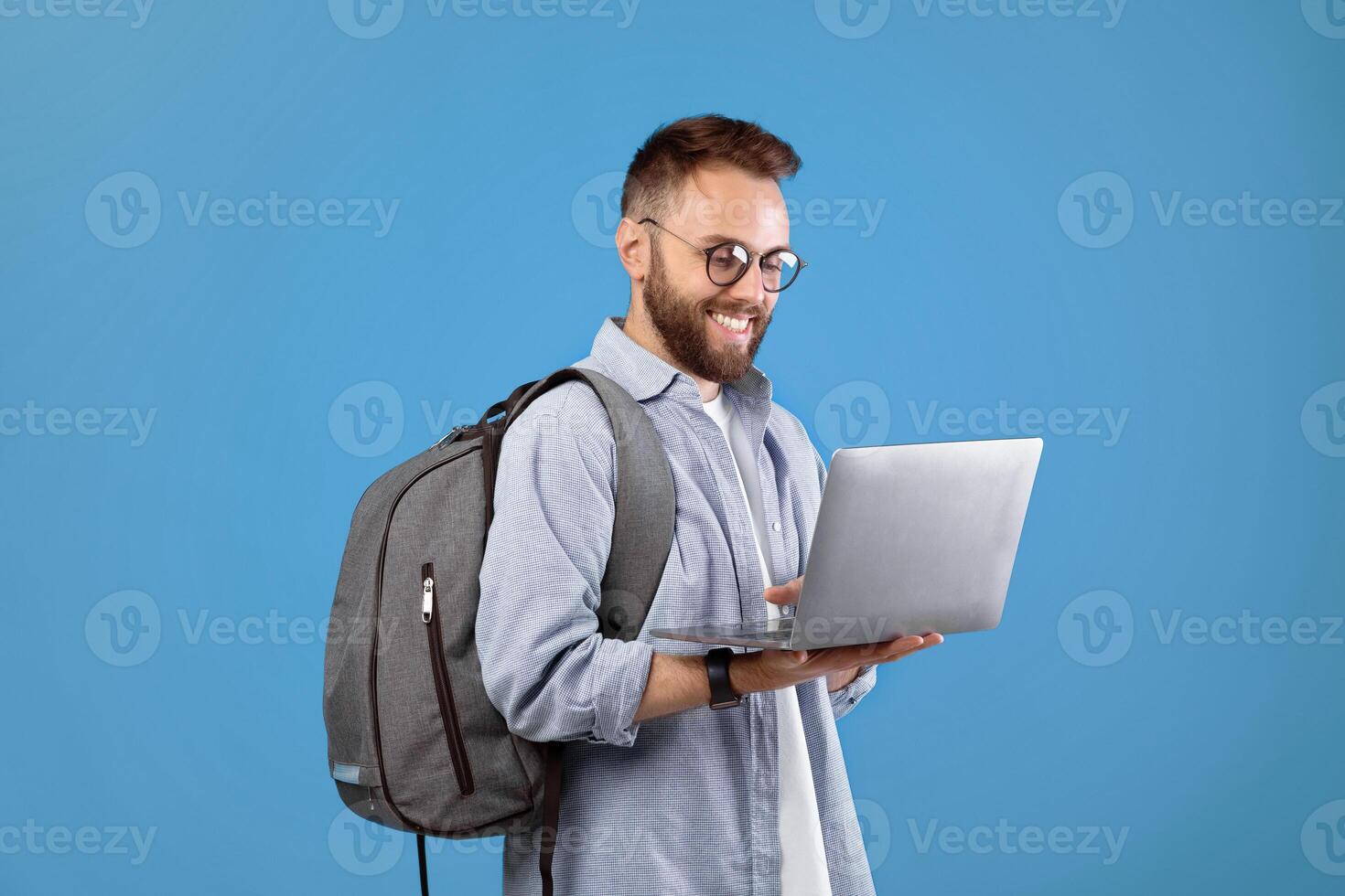 Cool millennial guy with backpack using laptop for online work or studies over blue studio background. Happy young man communicating on web, watching movie, browsing internet photo