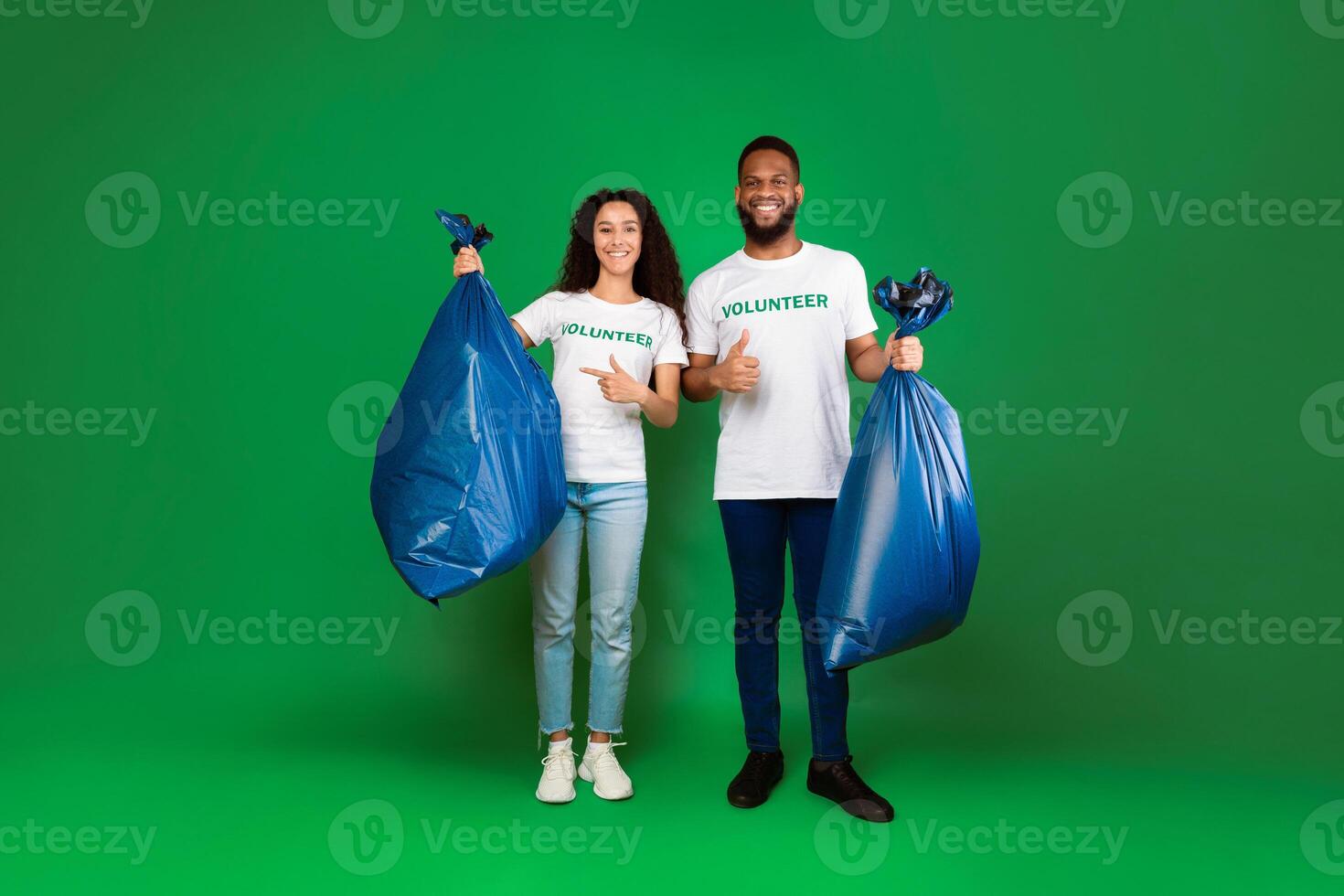 Keep Planet Clean. Two Diverse Volunteers Holding Blue Garbage Bags Picking Used Plastic Litter, Waste And Trash Standing On Green Studio Background. Volunteering Work, Environment Concept photo