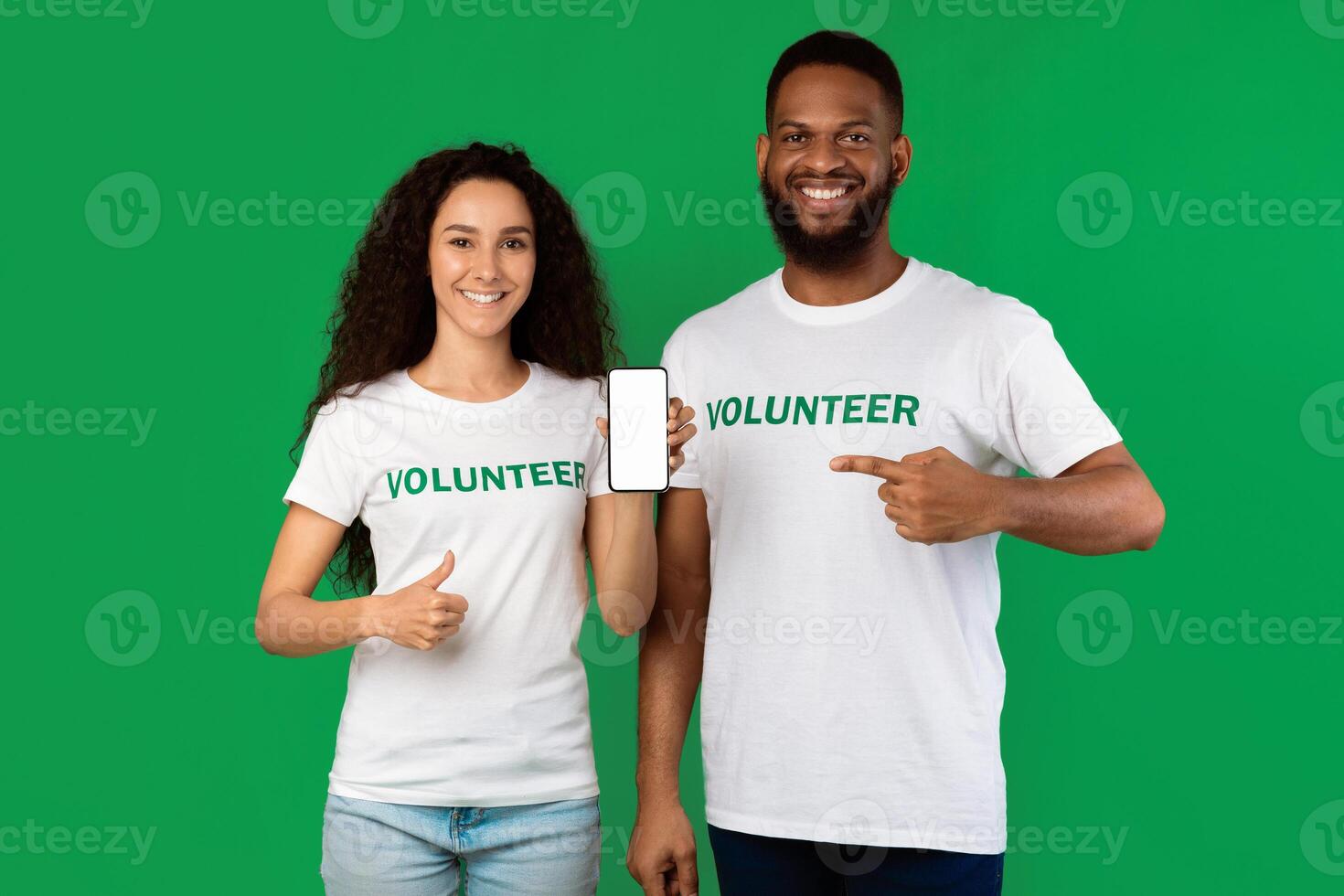 Female And Male Volunteers Showing Cellphone Blank Screen Gesturing Thumbs Up And Pointing Finger On Phone Standing On Green Studio Background, Smiling To Camera. Charity App Concept. Mockup photo