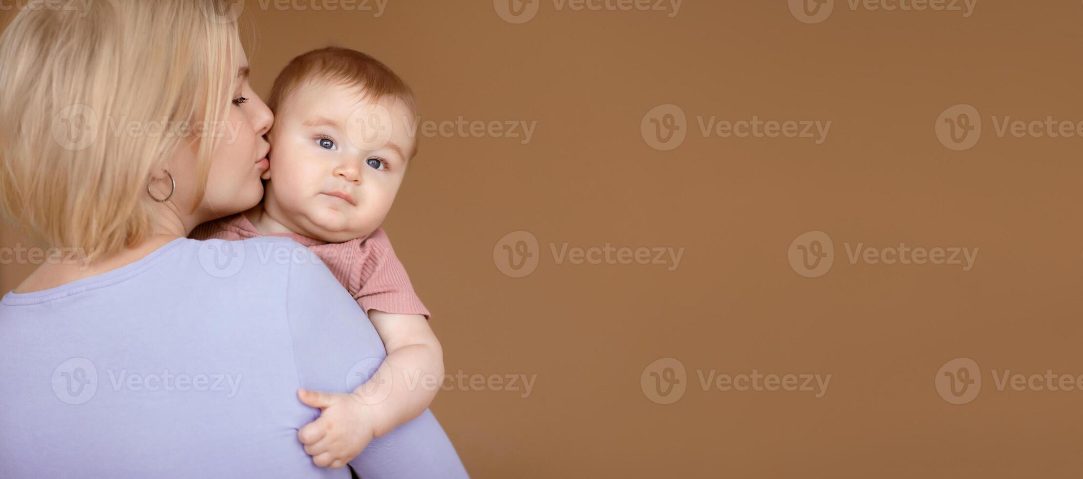 Closeup portrait of loving mother young blonde woman kissing little baby, brown studio background. Beautiful lady bonding with her adorable baby, holding cute sun or daughter and kissing, copy space photo