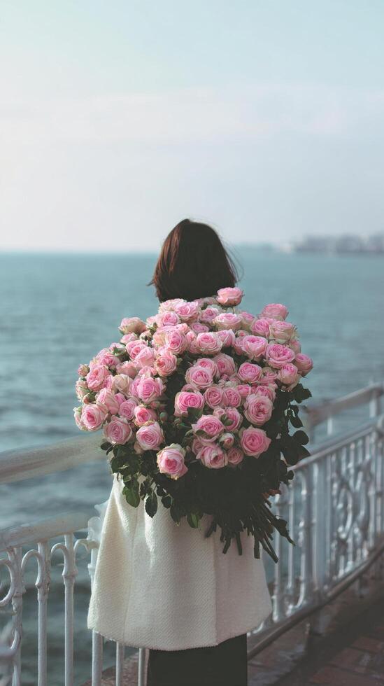 Woman Holding a Large Bouquet of Pink Roses While Standing by the Sea During a Calm Day photo