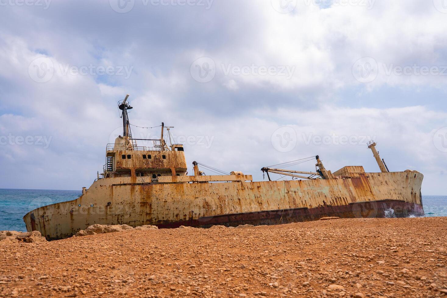 The Edro III shipwreck rests near Cyprus' rocky coast, its rusted hull contrasting with the vibrant blue sea and cloudy sky in a dramatic scene. photo