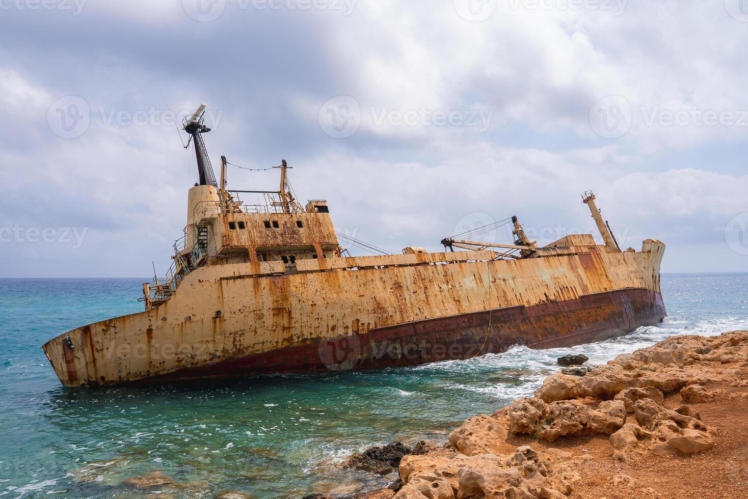 The Edro III shipwreck rests near the rocky coastline of Paphos, Cyprus, surrounded by turquoise waters and a partly cloudy sky, creating a dramatic scene. photo