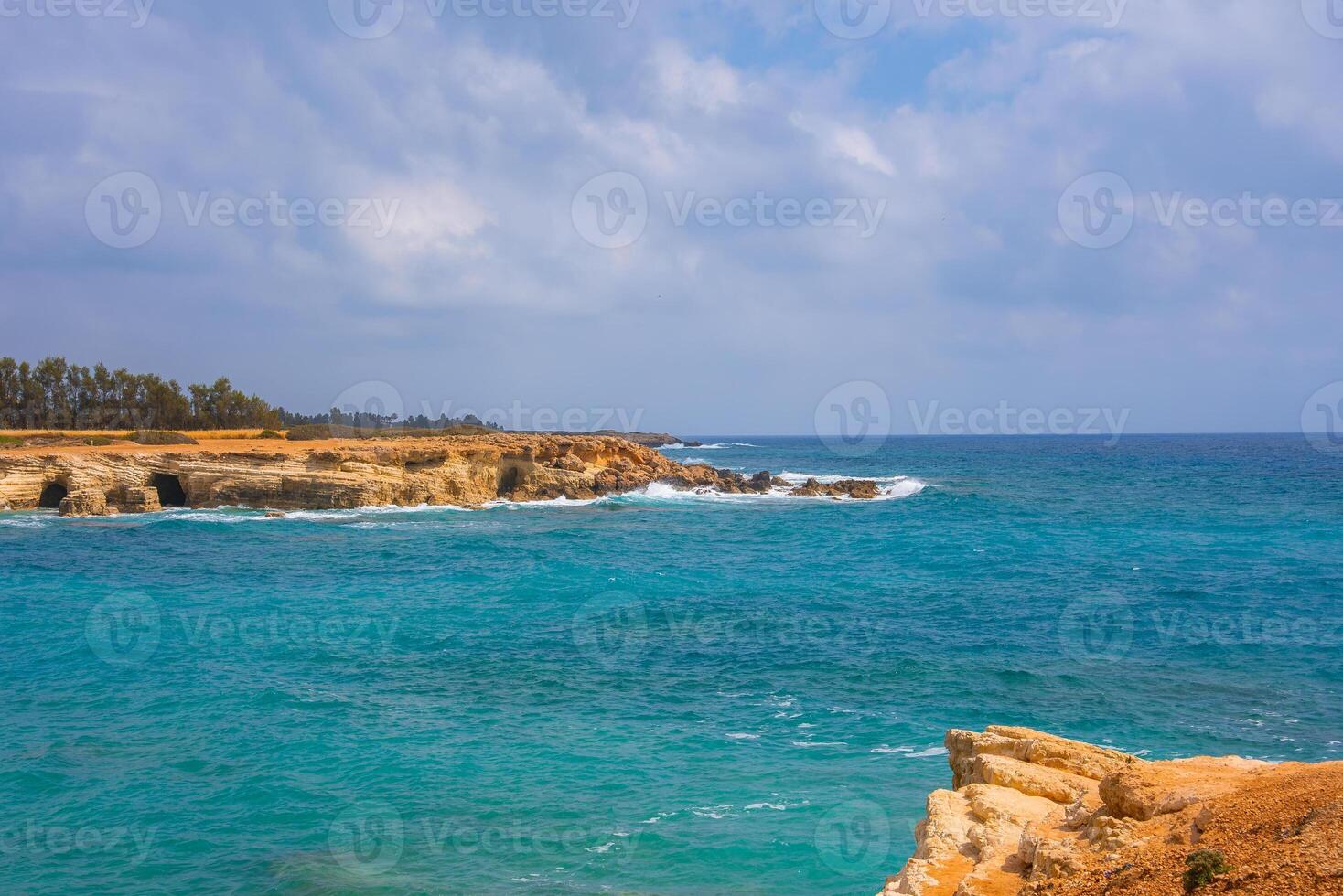 Coastal view of turquoise waters meeting golden cliffs with small caves, rocky formations, and a tree lined horizon under a partly cloudy sky. photo