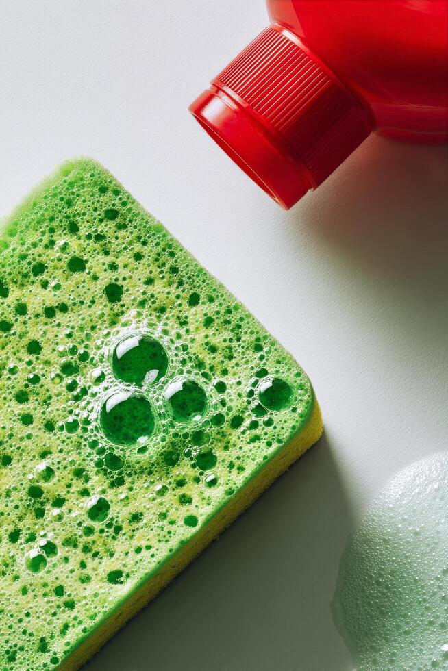 Bright Green Sponge and Red Soap Bottle With Bubbles on a White Surface During a Cleaning Session. photo