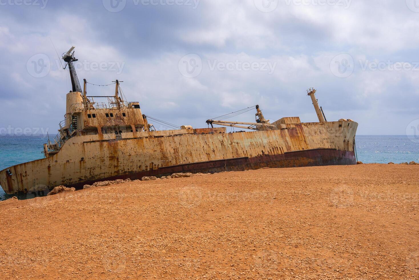 The Edro III shipwreck rests on a rocky coastline near Paphos, Cyprus, with its rusted hull leaning against a vibrant blue sea under a cloudy sky. photo