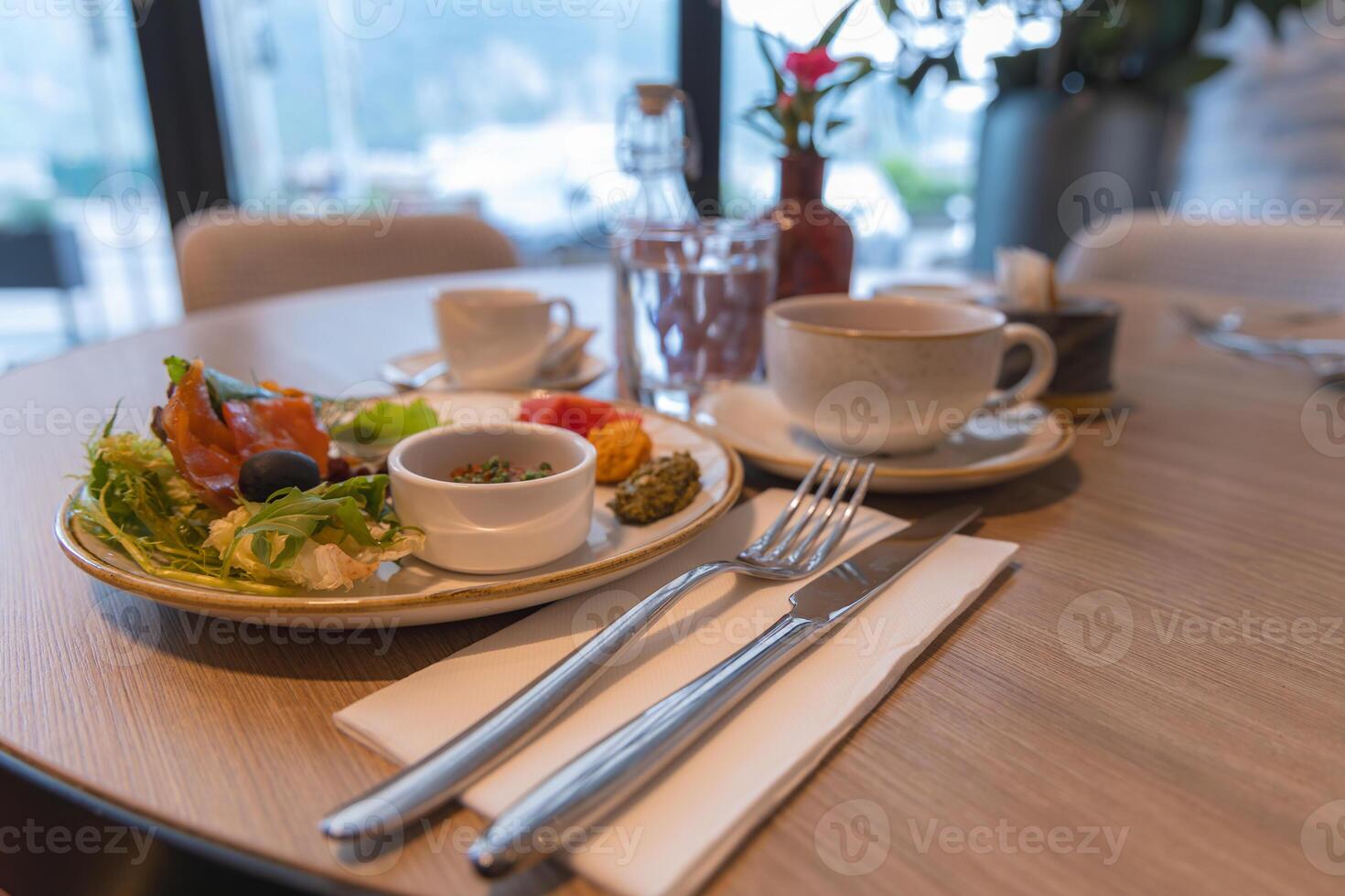 A dining table set with salad, smoked salmon, olives, coffee, and water. Adorned with fine cutlery and a red flower, with serene outdoor views. photo