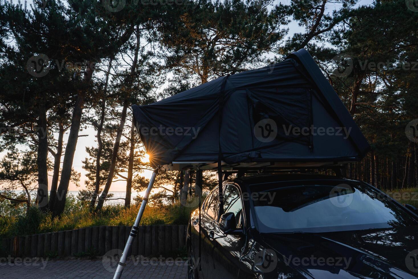 A modern SUV with a dark rooftop tent and ladder is parked in a forest. Tall trees surround the vehicle, with sunlight glowing through the canopy. A body of water is visible in the distance. photo