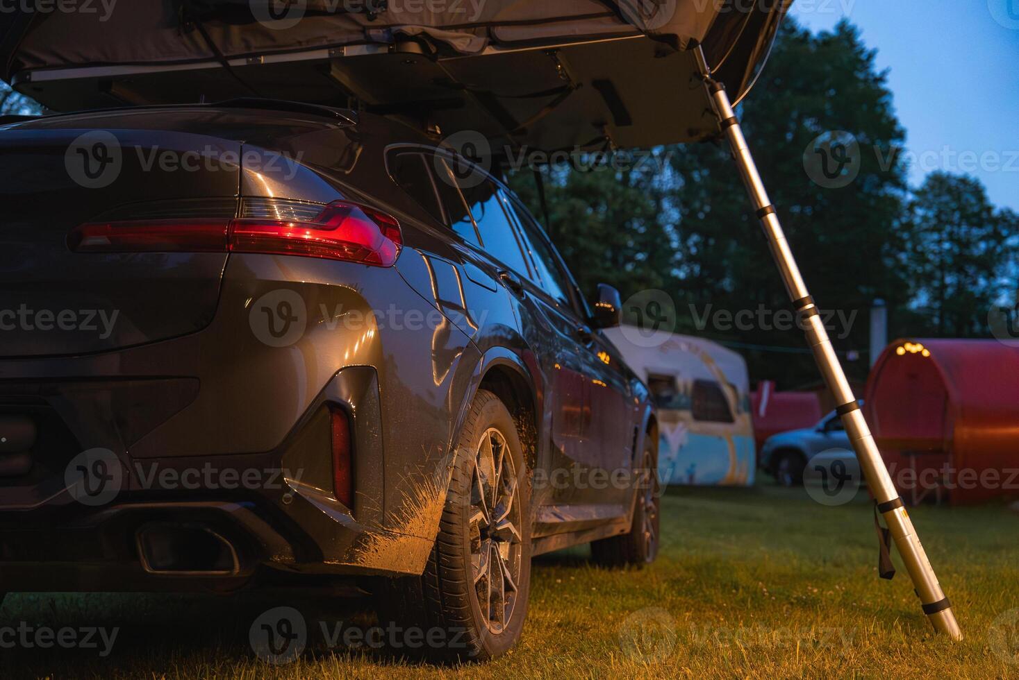A modern SUV with a foldable rooftop tent parked on grass in a forested campsite. Muddy details, camper trailers, and trees enhance the outdoor scene. photo
