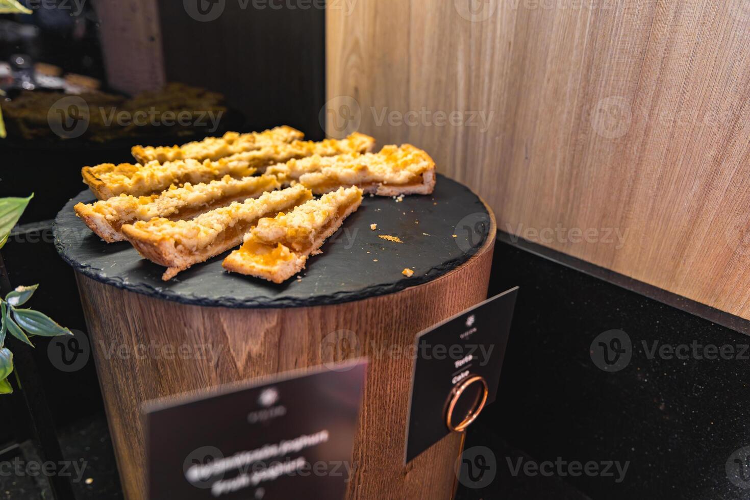 Display of sliced pastries with crumbly topping on a black slate platter atop a wooden stand, set against a wooden panel and black countertop. photo