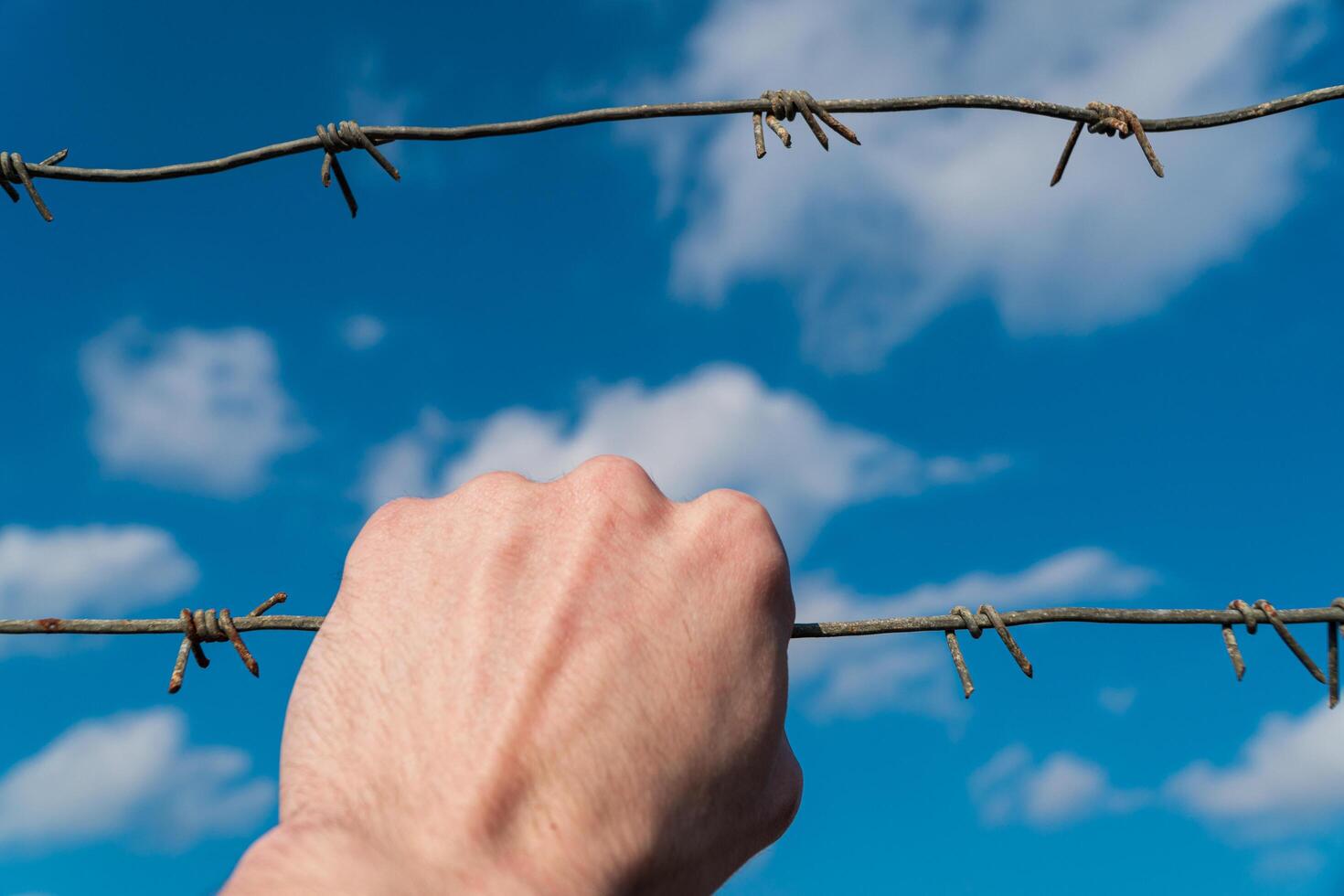 Man's hand holds stretched rusty barbed wire against blue sky with clouds. The concept of prison, crime, safety, border photo