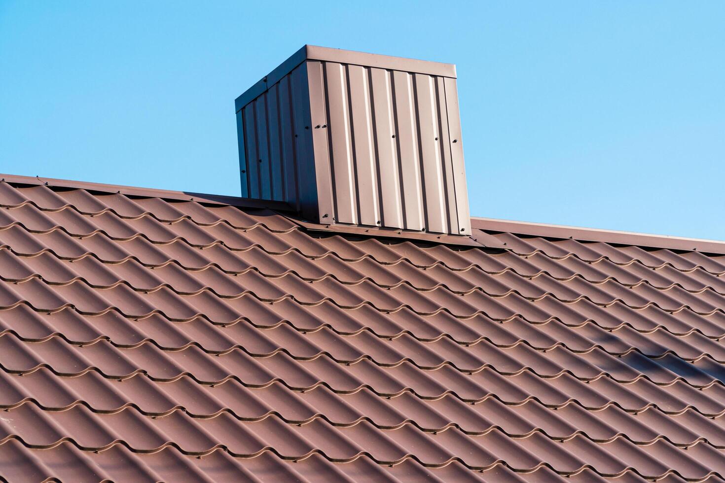 The roof of a house with modern metal tiles and a chimney, against the background of a blue sky. Metal roofing photo