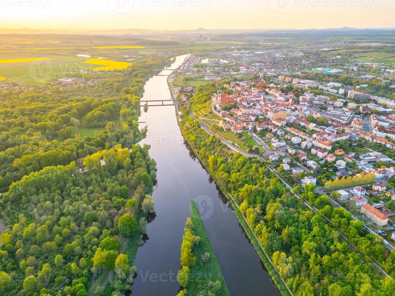 An aerial view captures the confluence of the Vltava and Labe rivers near Melnik, Czechia. The landscape features lush greenery, flowing waters, and a nearby town nestled against the rivers. photo