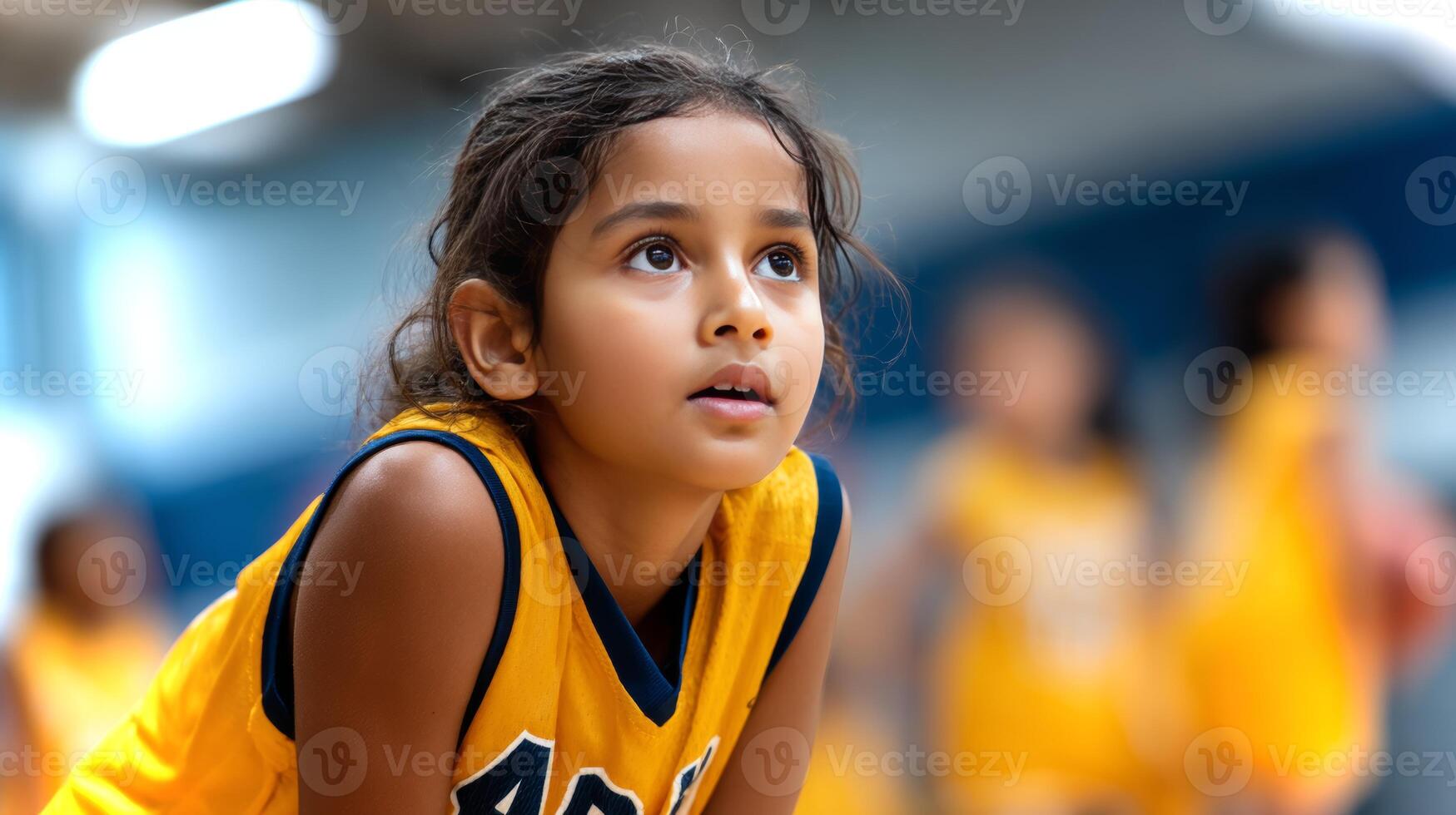 A young South Asian girl focuses intensely while in a defensive position during basketball practice. The gym is bustling with other players. photo