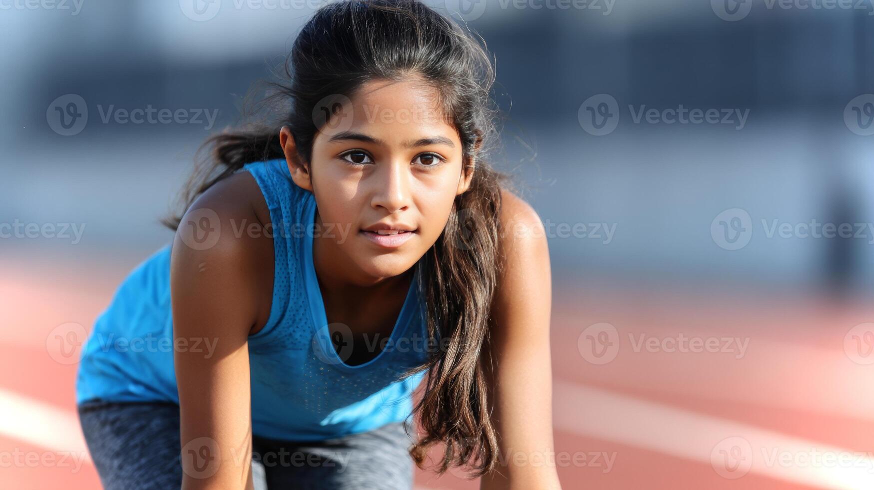 A South Asian girl kneels in a start pose on a red running track, showing concentration and readiness to sprint. photo