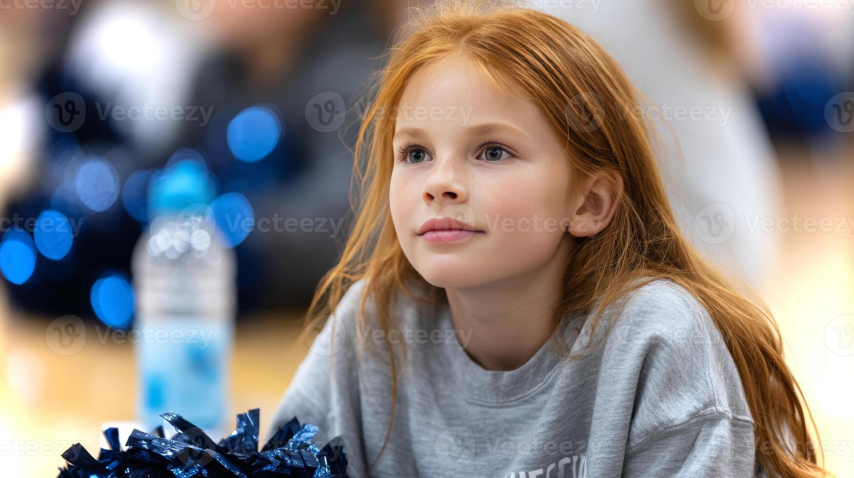 A red-haired girl enjoys a restful moment between cheerleading practice, sitting in a sports hall surrounded by teammates. She appears focused and relaxed. photo