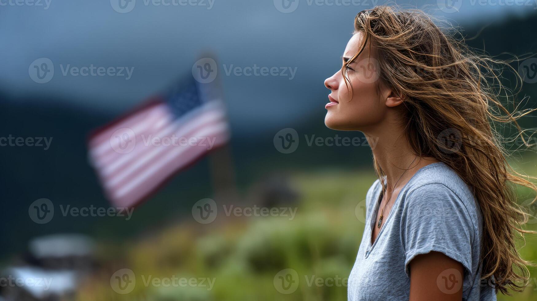 A girl with long hair stands at the edge of a lush green field, gazing thoughtfully as a flag flutters behind her on a cloudy day. photo