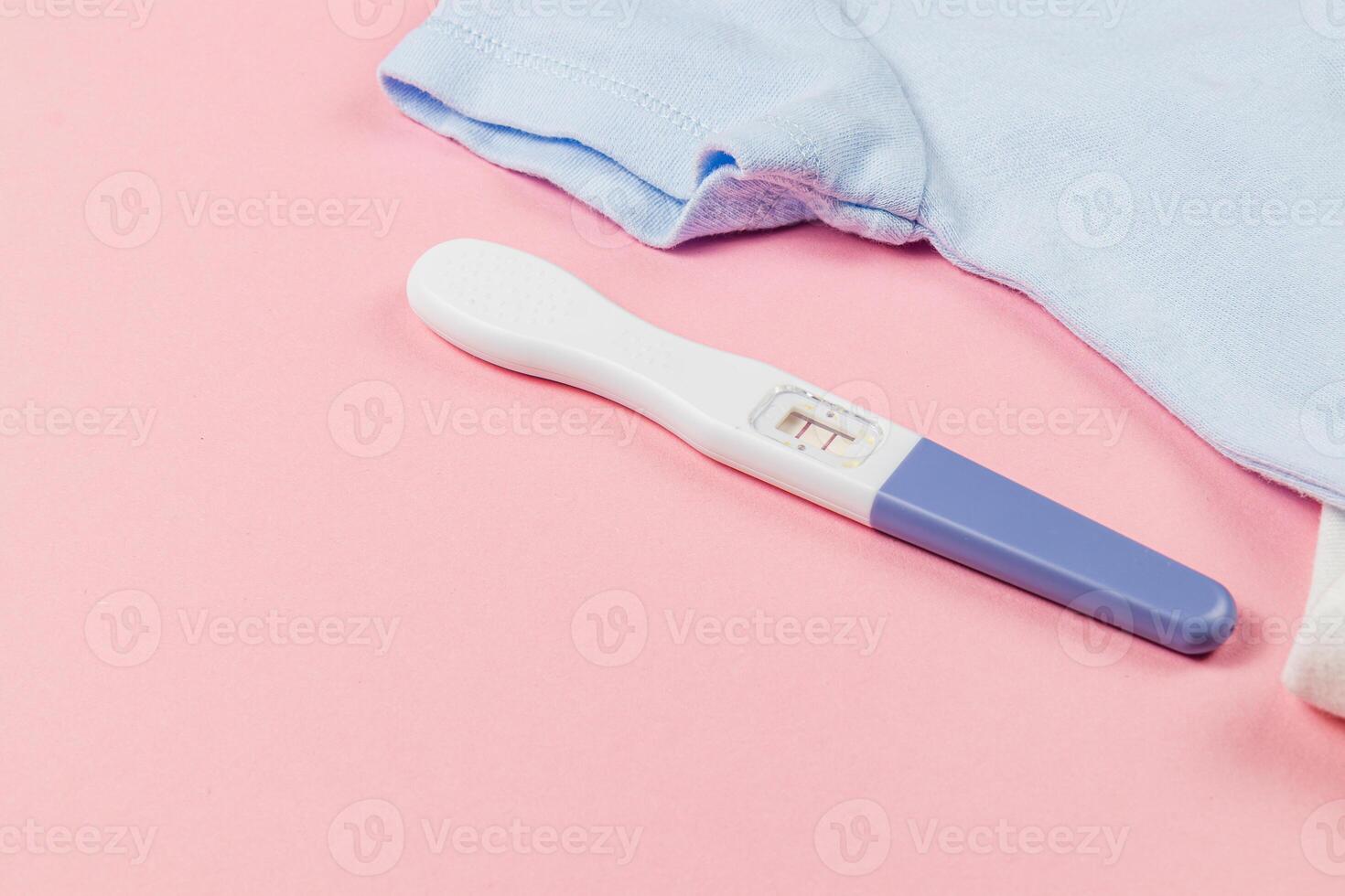 Positive pregnancy test next to baby clothes on pink background photo
