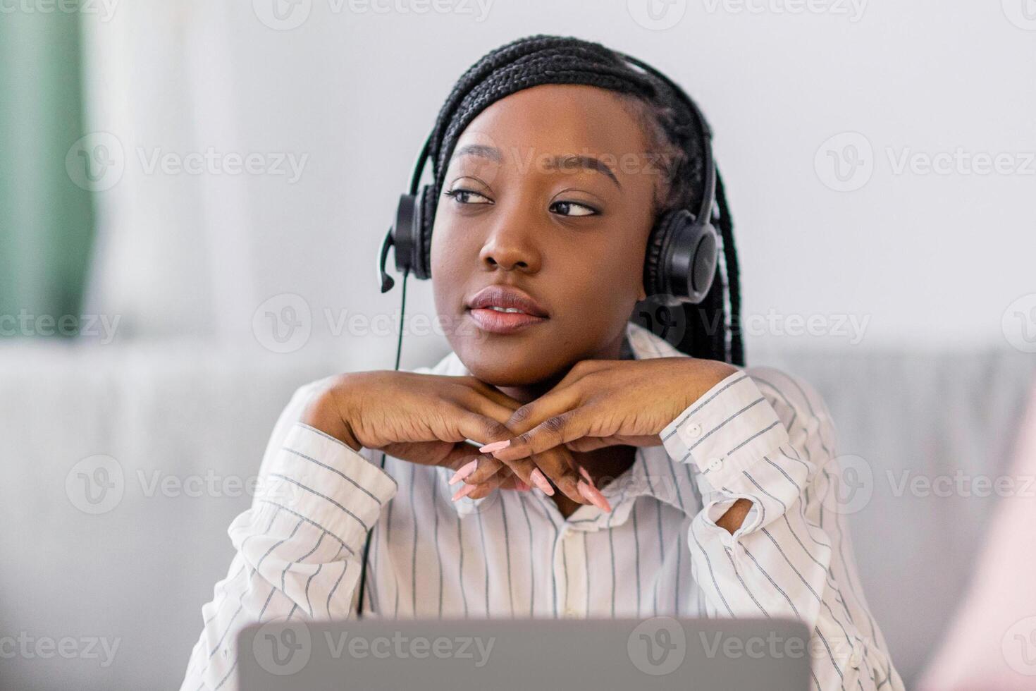 Pensive young black lady having online training at home, sitting at workdesk in front of laptop, leaning on her elbows and looking at copy space, using headphones. E-education, online class concept photo