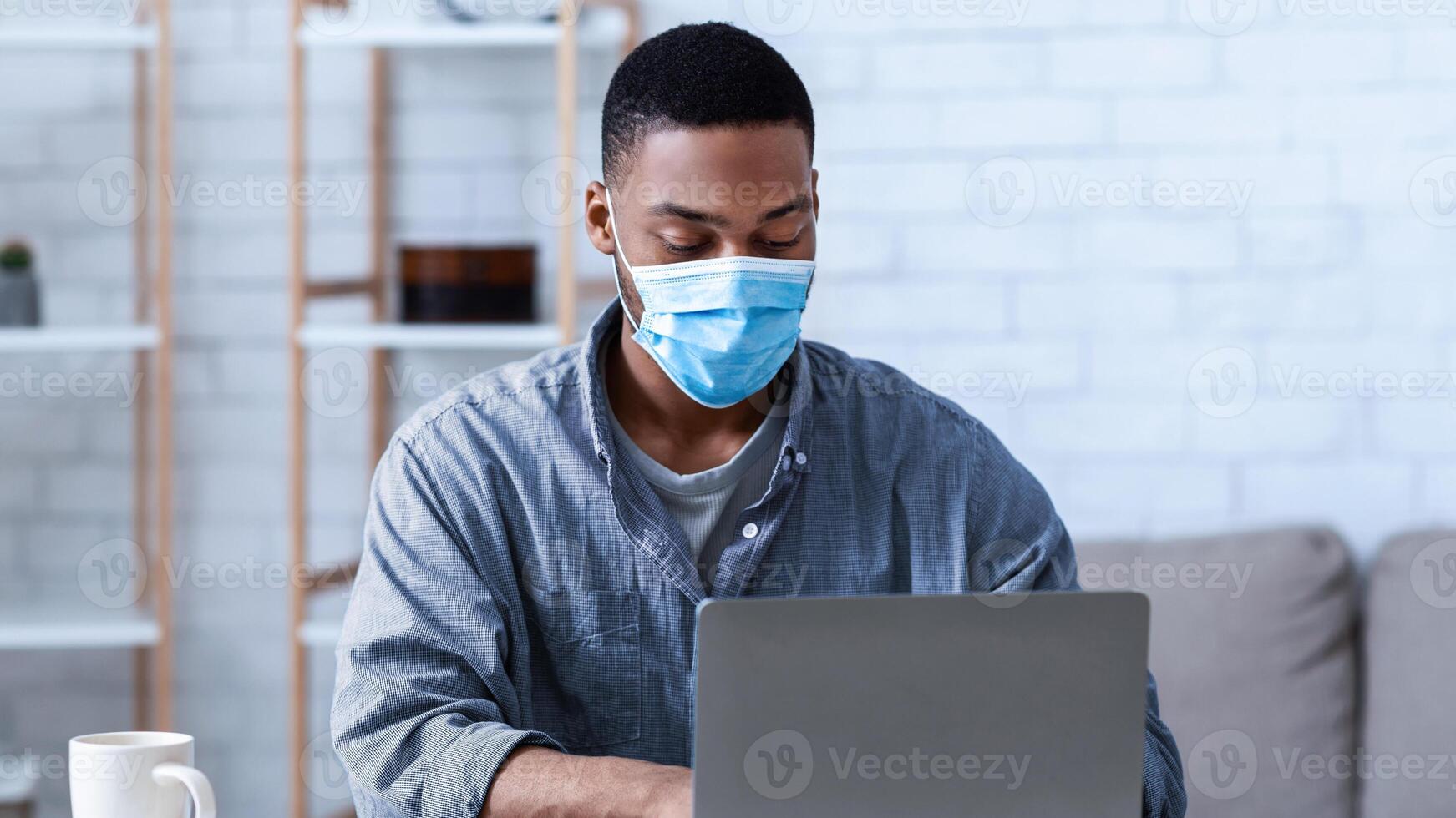 Black Man Wearing Face Mask Working On Laptop Sitting In Modern Office. Social Distancing At Work During Coronavirus Covid-19 Pandemic. Remote Work Concept. Selective Focus photo