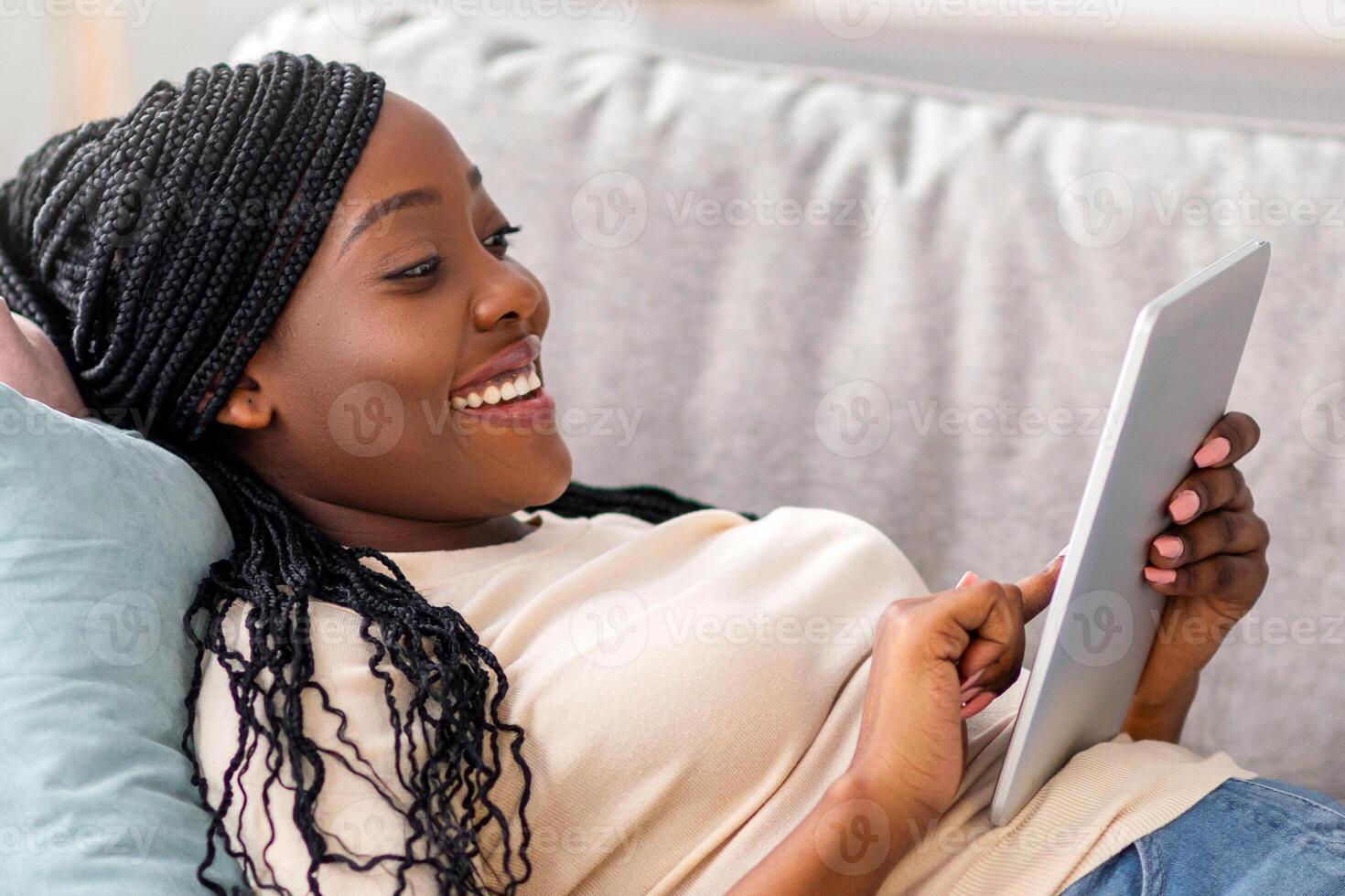 Joyful black woman watching movie, using mobile application on pad. Relaxed african american young lady laying on couch, using digital tablet and eating popcorn, home interior, copy space photo