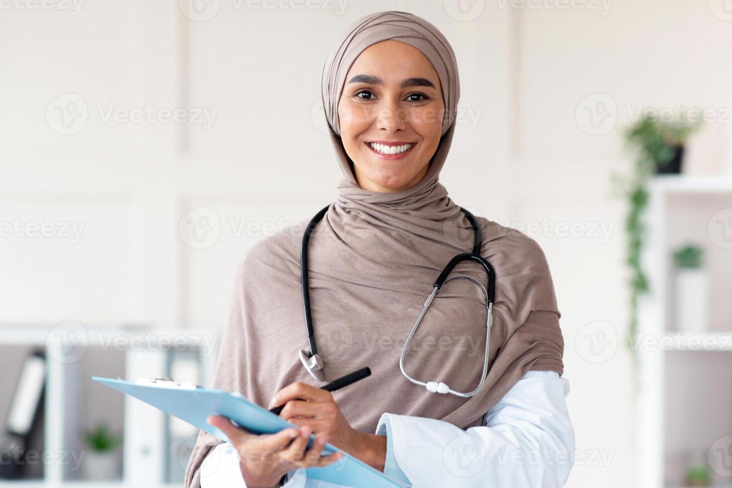 Cheerful pretty woman in hijab doctor posing in cabinet, holding medical folder and pen, having stethoscope, copy space. Happy muslim lady greeting patients at hospital, smiling at camera photo