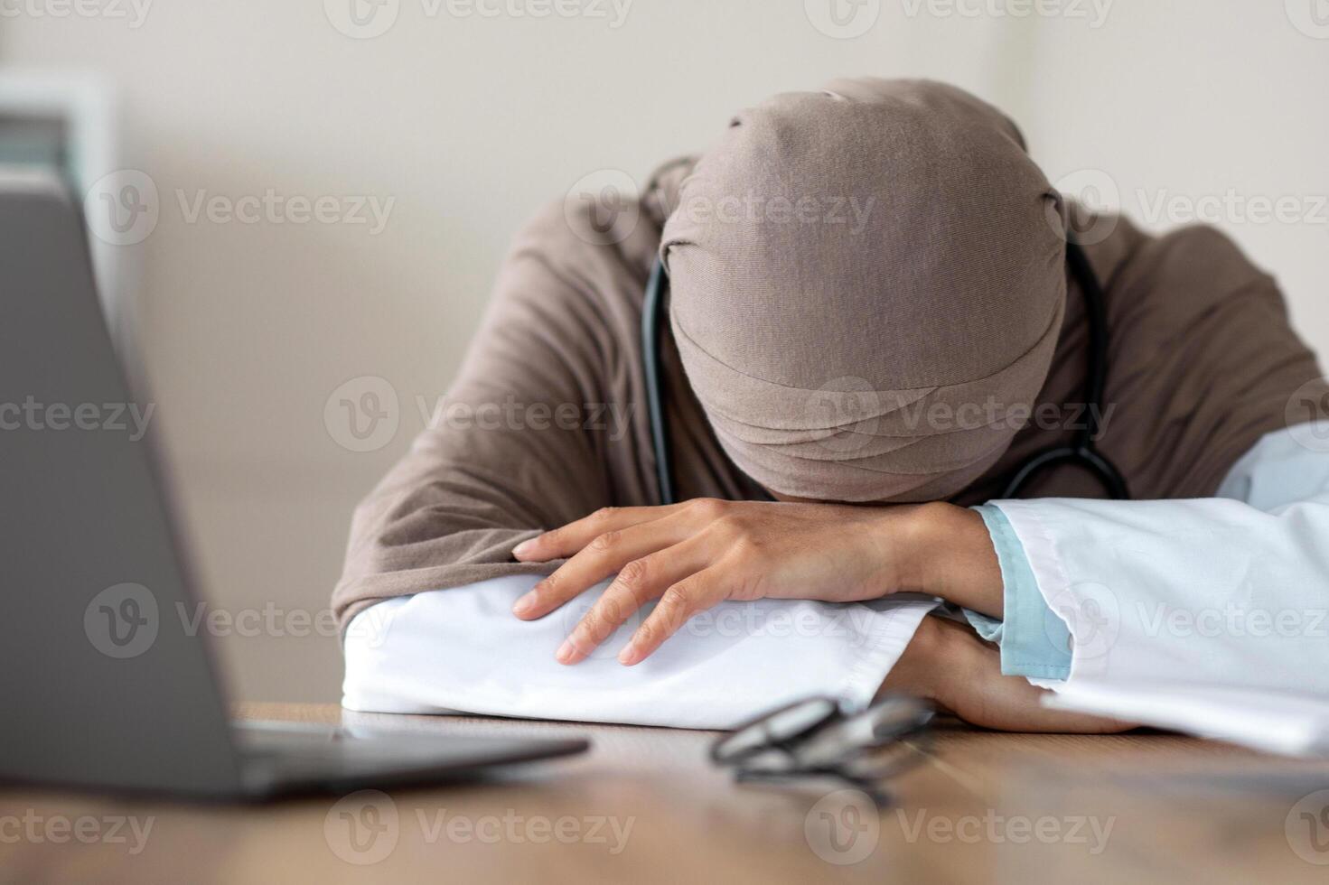 Overworked female doctor woman in hijab having nap at workdesk, working with laptop in clinic. Exhausted physician young lady leaning on her hands, needs for break during long working day photo