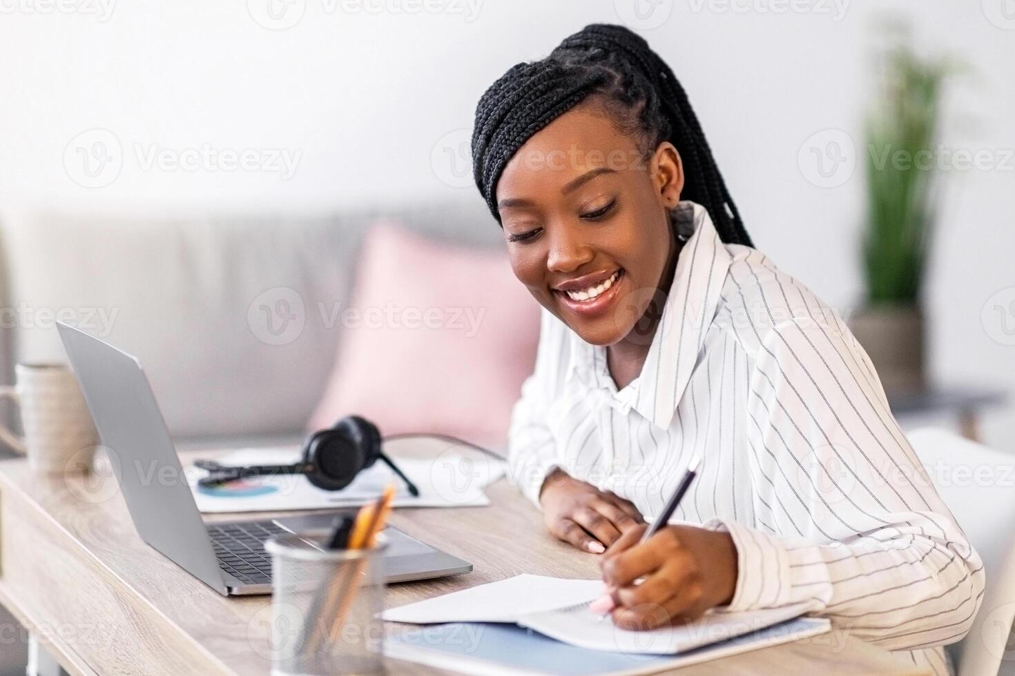 Pretty black woman student studying in front of laptop, taking notes and smiling, side view, copy space. Cheerful african american young lady attending webinar while staying home during lockdown photo