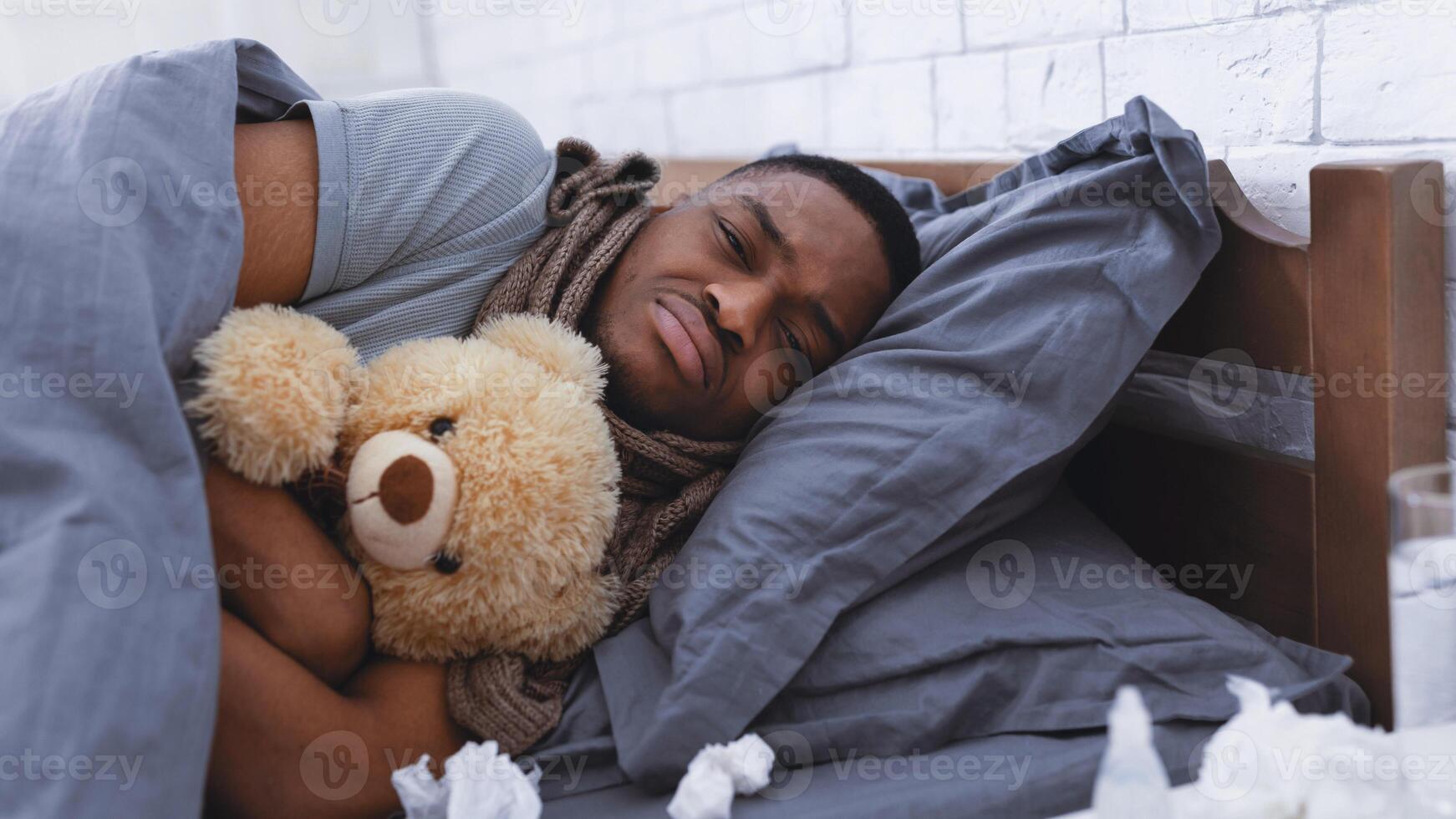 Depressed Sick African Man Hugging Teddy Bear Toy Lying Wrapped In Blanket Having Cold In Bedroom At Home. Flu And Depression, Mental Health During Coronavirus Disease Concept. Selective Focus photo