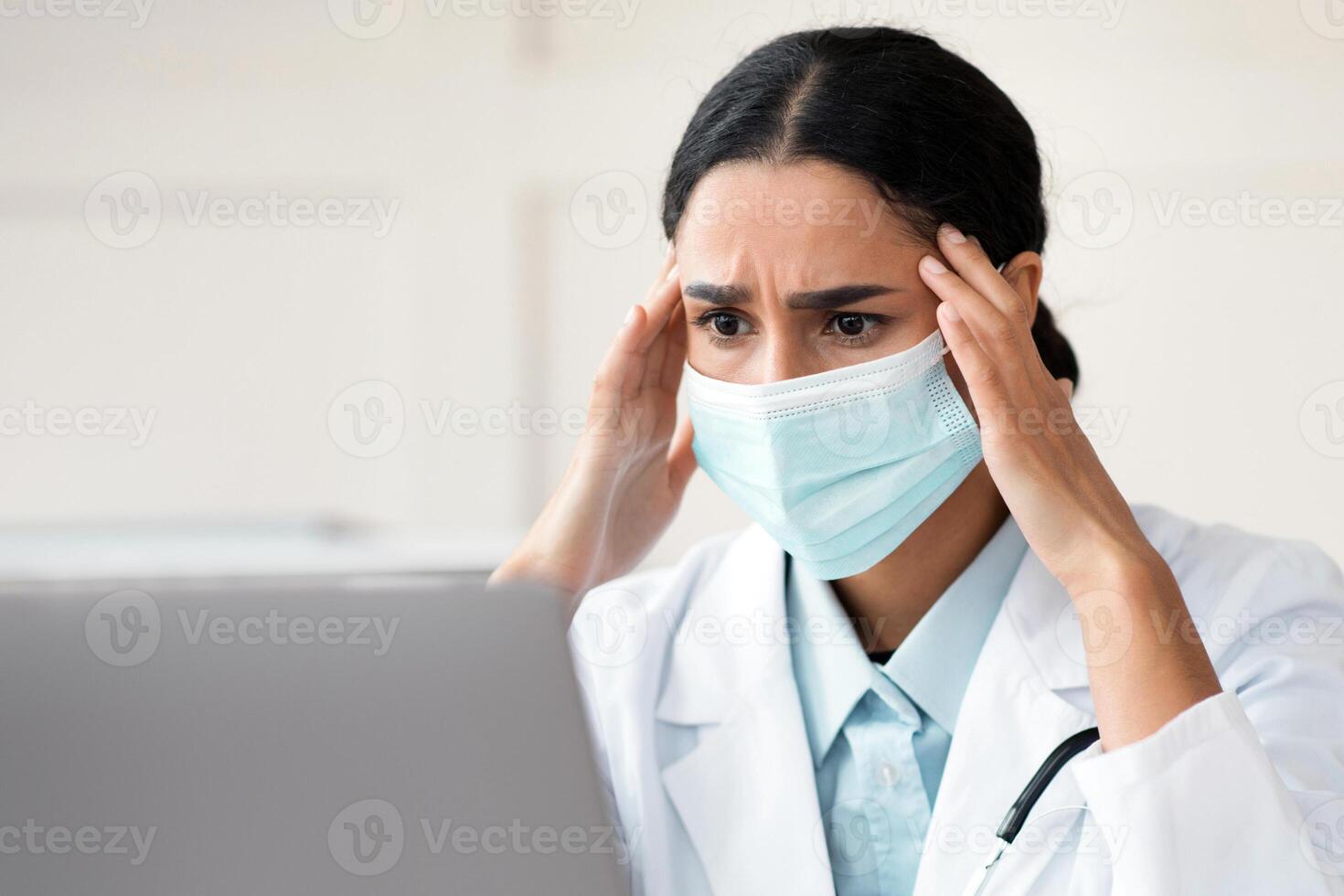 Exhausted arab female doctor in protective face mask working in clinic, sitting at workdesk in front of laptop, touching her head, need for break, copy space. Brunette woman physician having headache photo