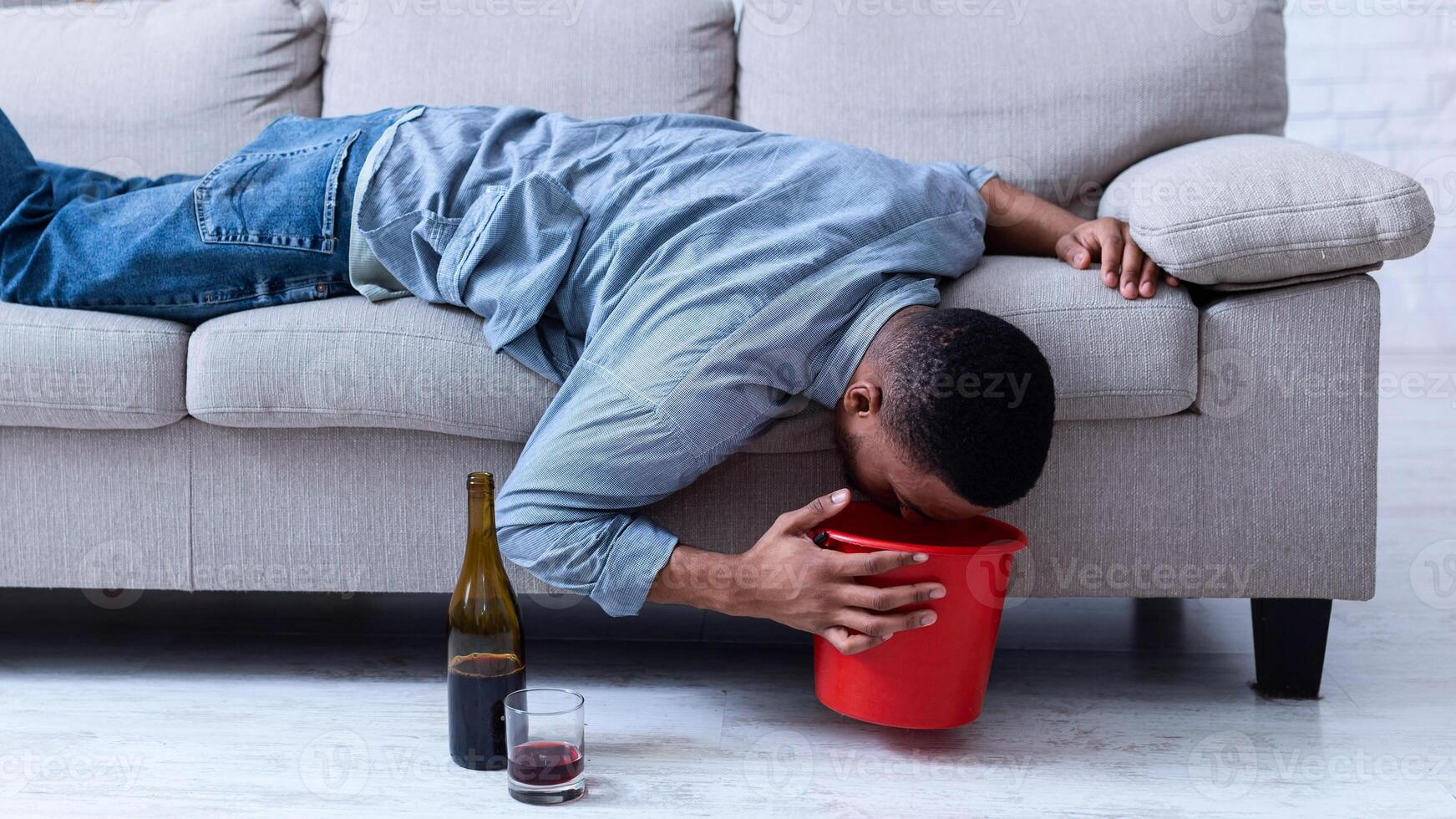 Alcohol Poisoning. Drunk Man Vomiting Into A Bucket After Drinking Alcoholic Wine Lying On Sofa At Home. Booze Intoxication, Alcoholism Addiction Habit Concept. photo