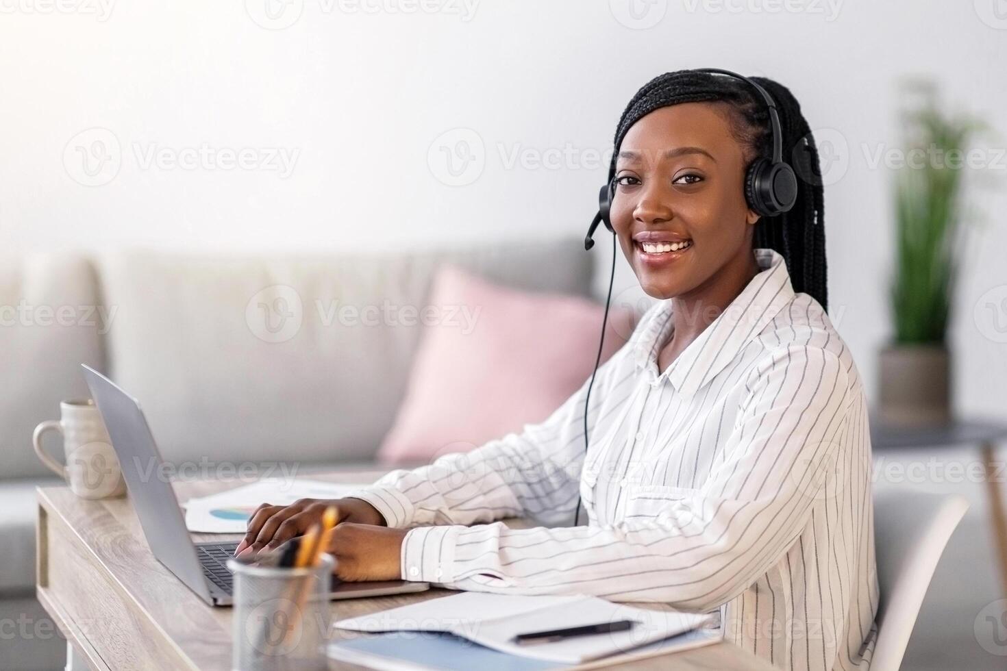 Pretty black woman smiling at camera while listening to webinar from home, using headset and laptop, side view, copy space. Cheerful african american lady working online, typing on computer keyboard photo