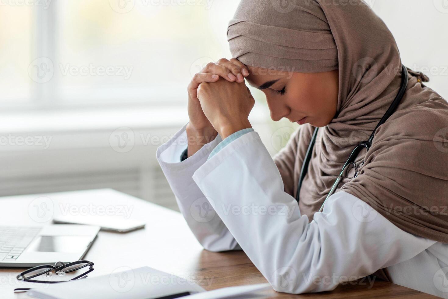 Side view of exhausted muslim woman doctor in hijab working in clinic, sitting at her workdesk in front of laptop, leaning on her hand, copy space. Tired lady in hijab physician having headache photo