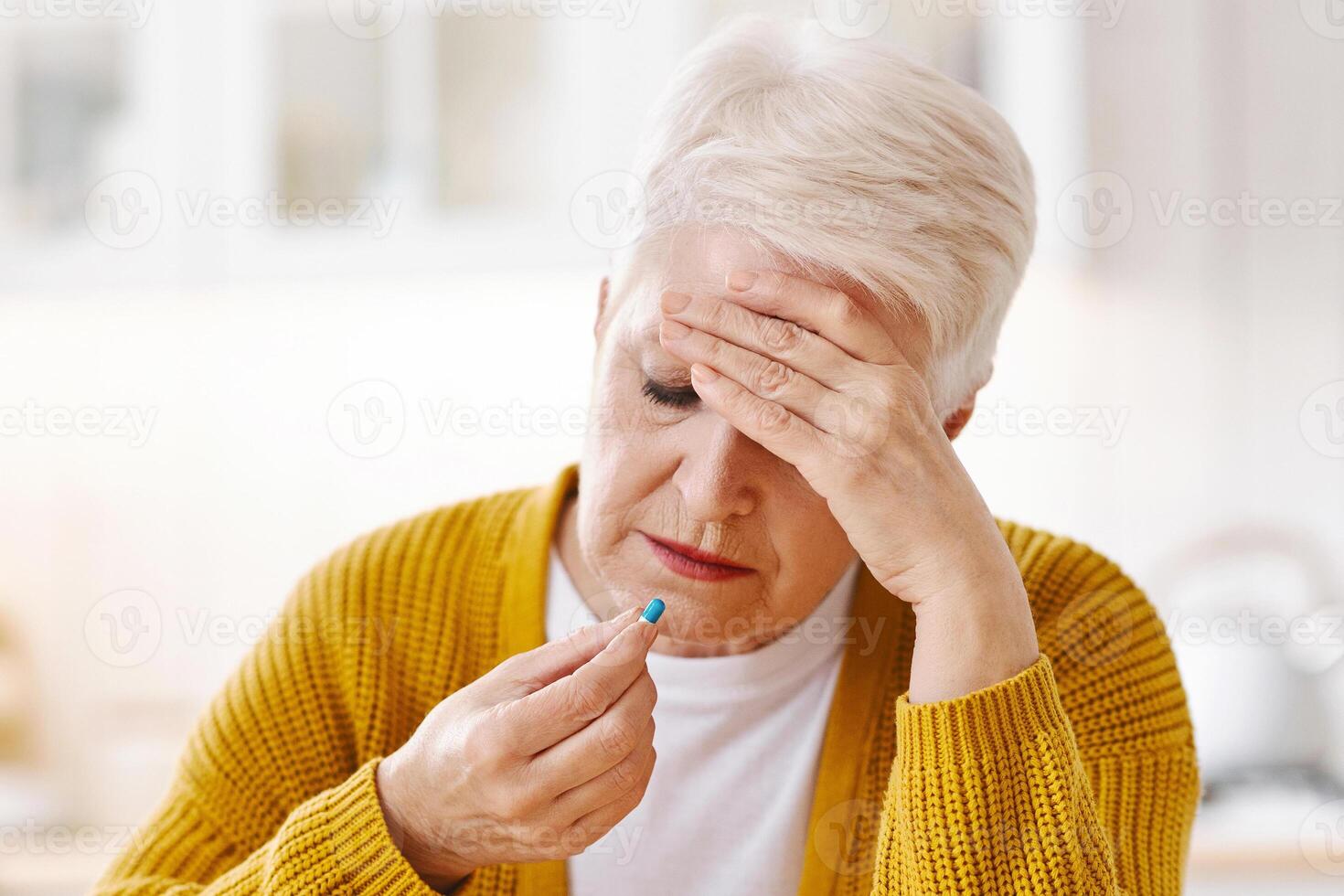 Upset old woman suffering from headache, touching her forehead and taking pill, kitchen interior, copy space. Grey-haired elderly lady feeling bad, having migraine or tension problem photo