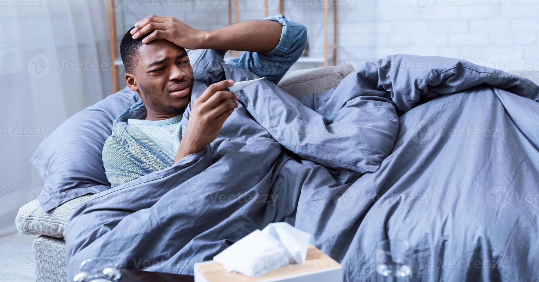 Sick African Man Having Fever Touching Forehead Holding Thermometer Lying On Couch At Home. Ill Black Guy Measuring Temperature. Influenza Symptoms Concept. Selective Focus photo