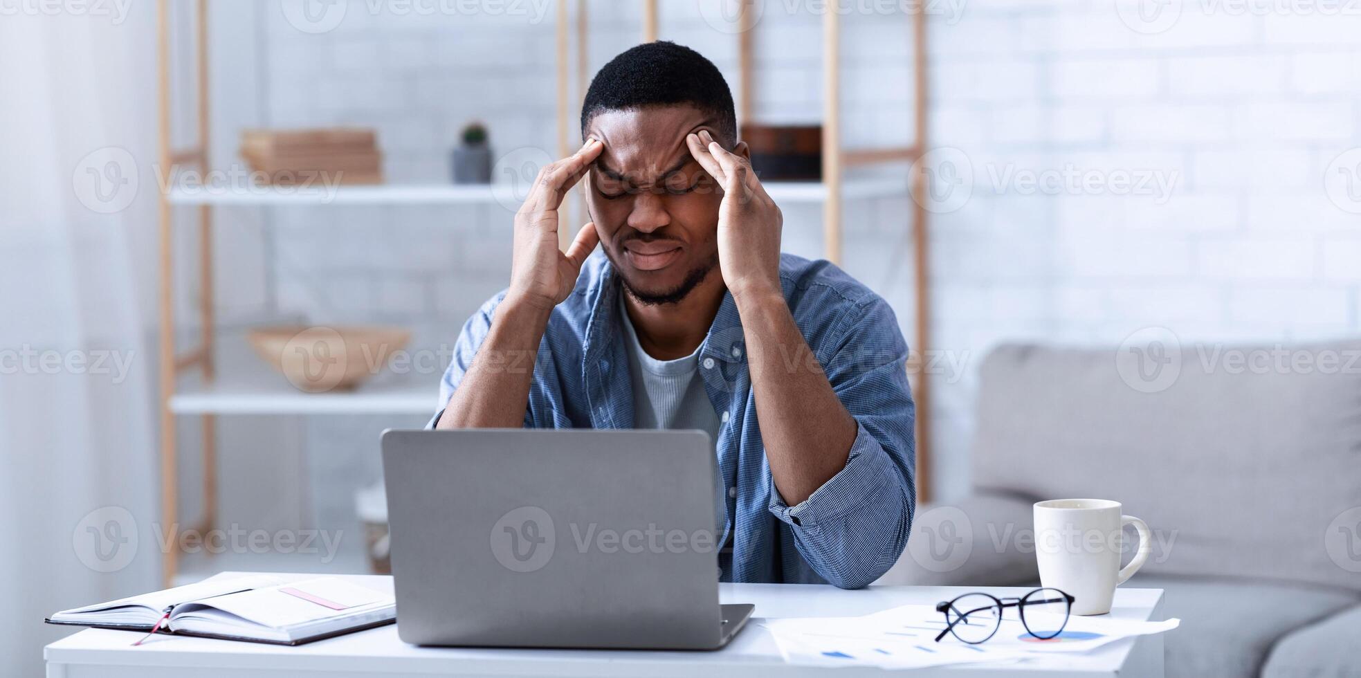 African American Businessman Having Headache Sitting At Laptop Computer Working From Home. Black Man Massaging Temples Suffering From Migraine Pain Indoor. Selective Focus photo