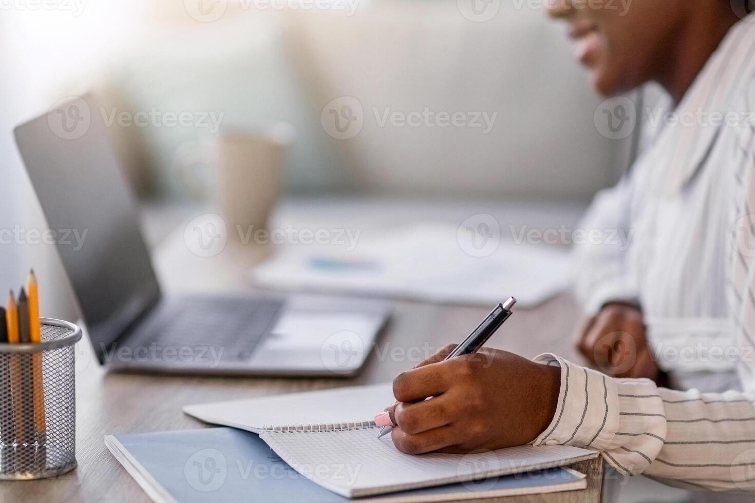 Closeup of young black woman having online course, smiling african american lady in headset sitting at table in front of laptop, taking notes in notepad, home interior, side view, copy space photo