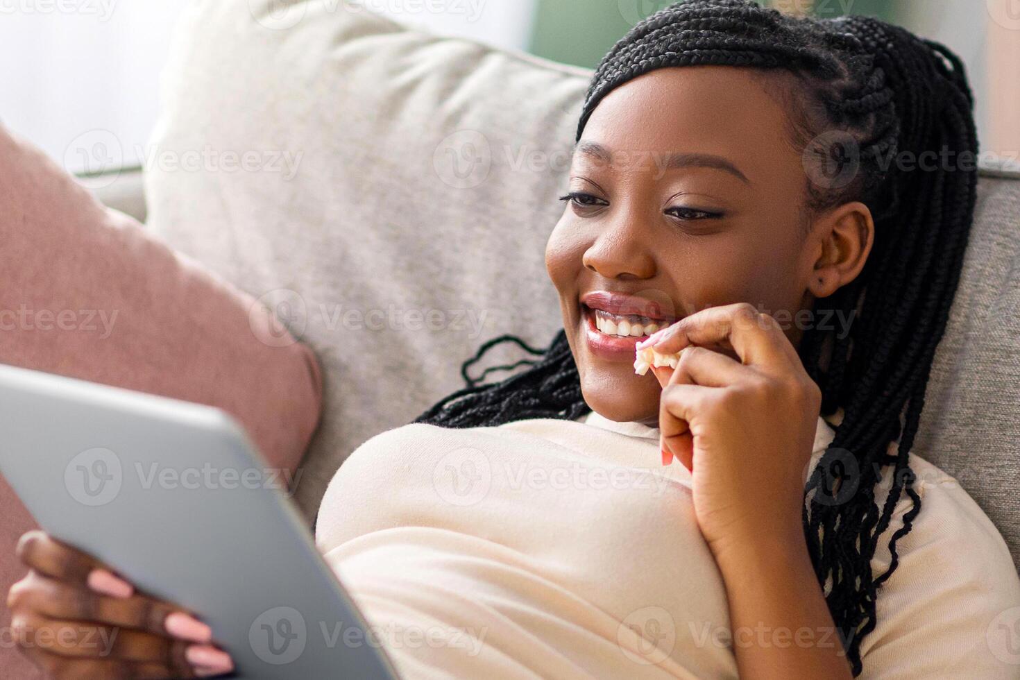 Relaxed african american young lady reclining on couch, using digital tablet and eating popcorn, home interior, copy space. Joyful black woman watching movie, using mobile app on pad photo
