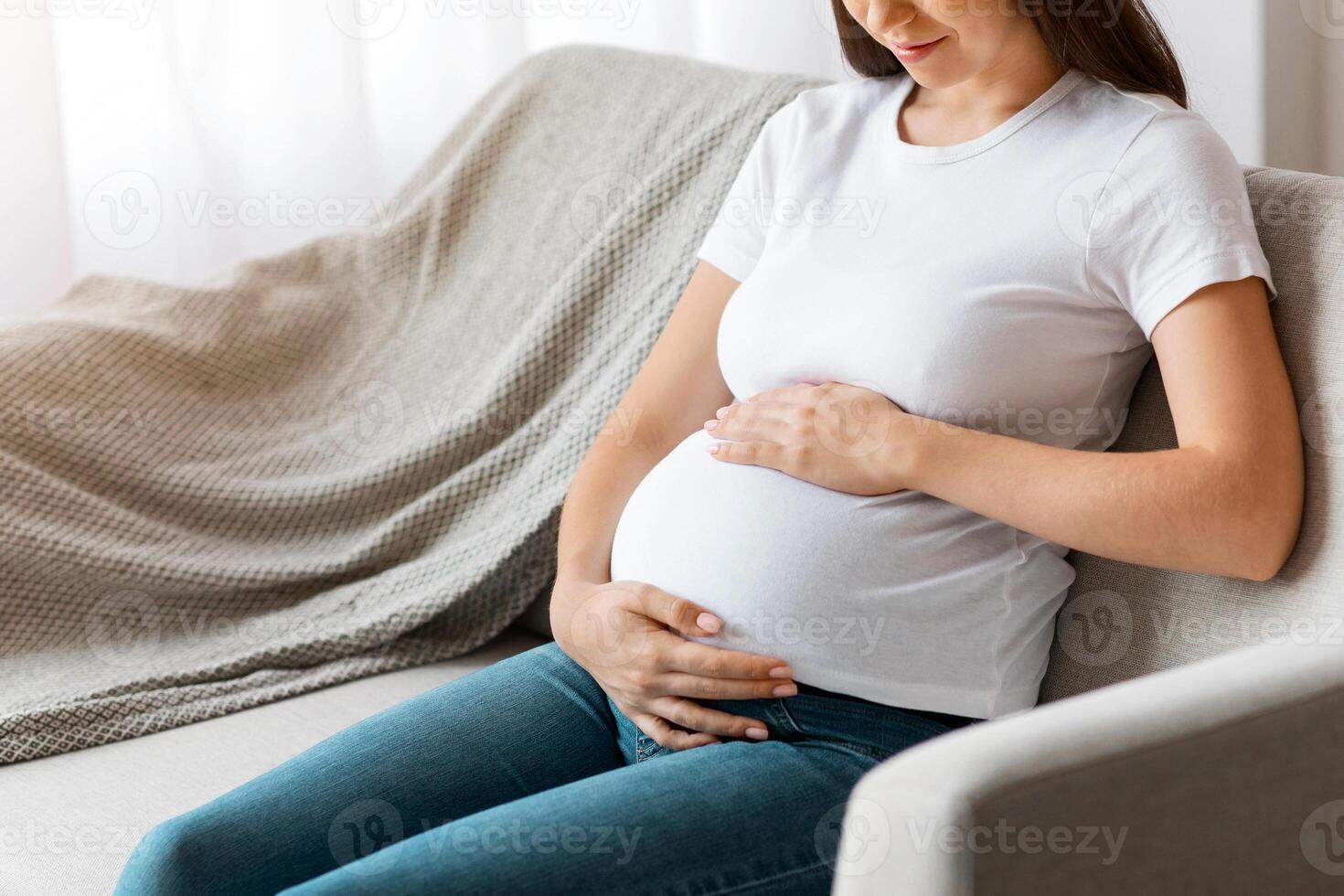 Maternity Concept. Young Beautiful Woman Expecting Baby Relaxing On Couch At Home, Happy Young Smiling Woman Tenderly Embracing Her Pregnant Belly While Resting On Comfortable Sofa, Copy Space photo