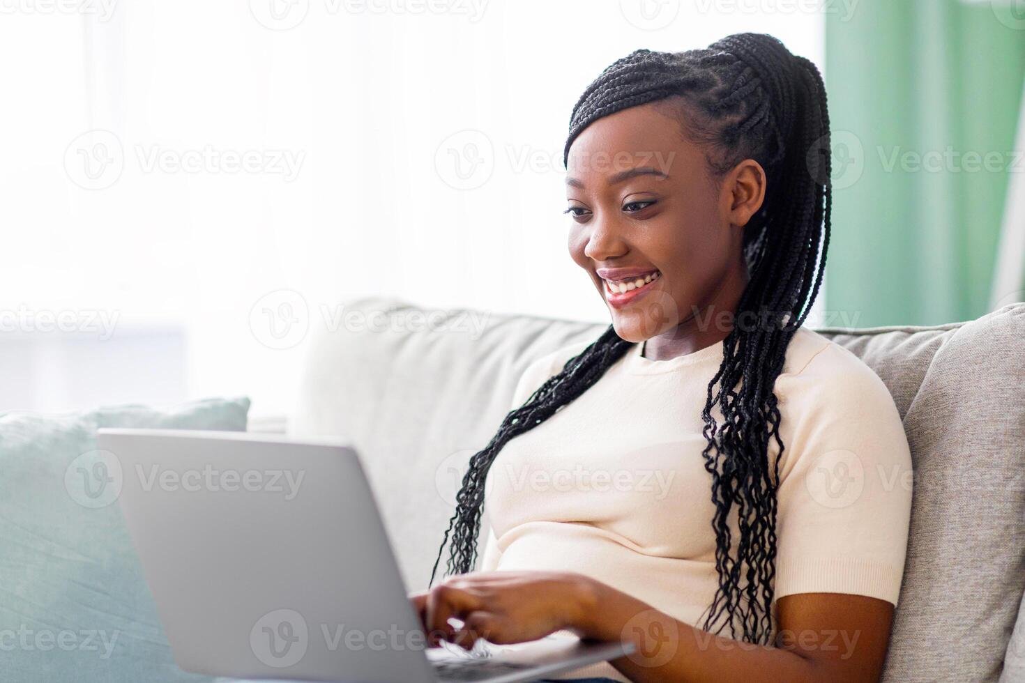 Happy young black woman with cute hair freelancer working from home, sitting on couch and using modern laptop, typing text or communicating with clients online, blogging or copywriting, copy space photo
