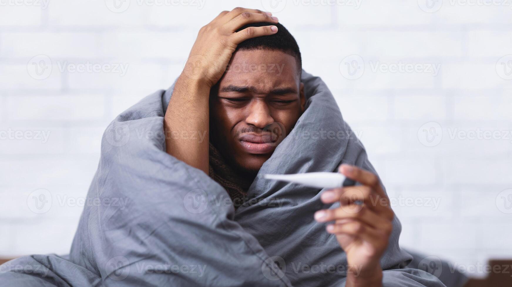 Fever. Portrait Of Sick African American Man With Thermometer Touching Aching Head Suffering From Flu Symptoms Sitting Wrapped In Blanket In Bed At Home. Selective Focus photo