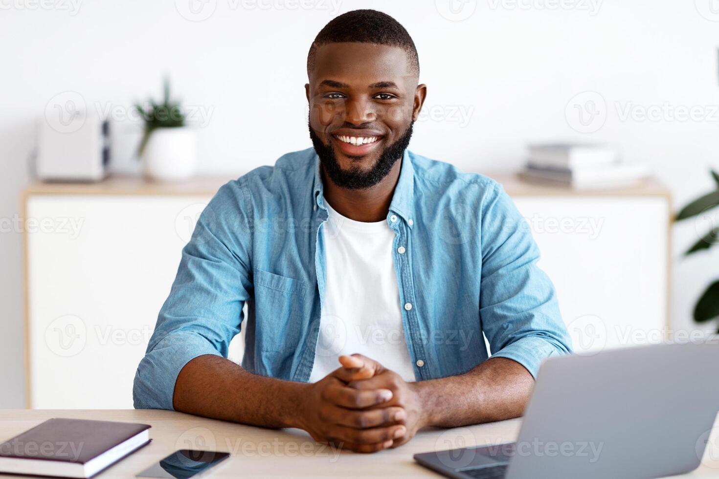 Portrait Of Young Black Freelancer Guy Sitting At Desk With Laptop Computer At Home Office, Looking At Camera, Smiling African American Entrepreneur Man Enjoying Remote Work And Self-Employment photo