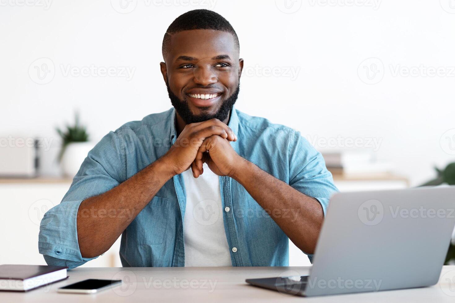 Pensive Black Man Office Worker Sitting At Desk With Laptop And Smiling. Handsome African American Freelancer Guy Thinking About Business Strategy At Workplace, Resting Chin On Hands And Looking Away photo