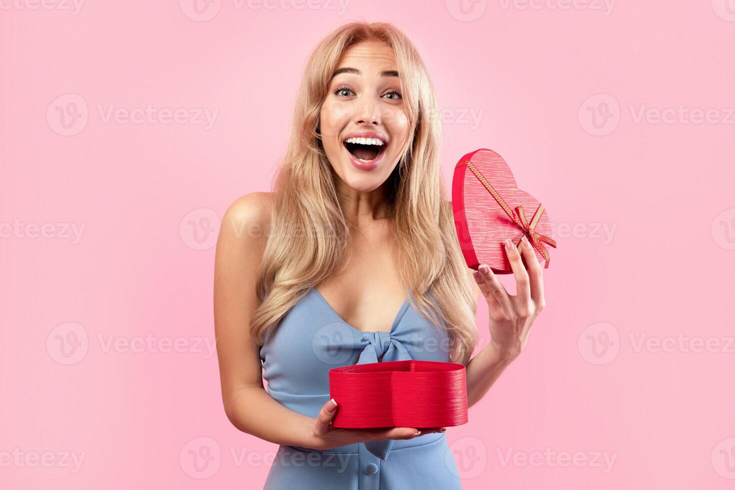 Excited blonde woman opening heart shaped gift box for Valentine's Day over pink studio background. Gorgeous millennial lady happy to receive present for lover's holiday photo