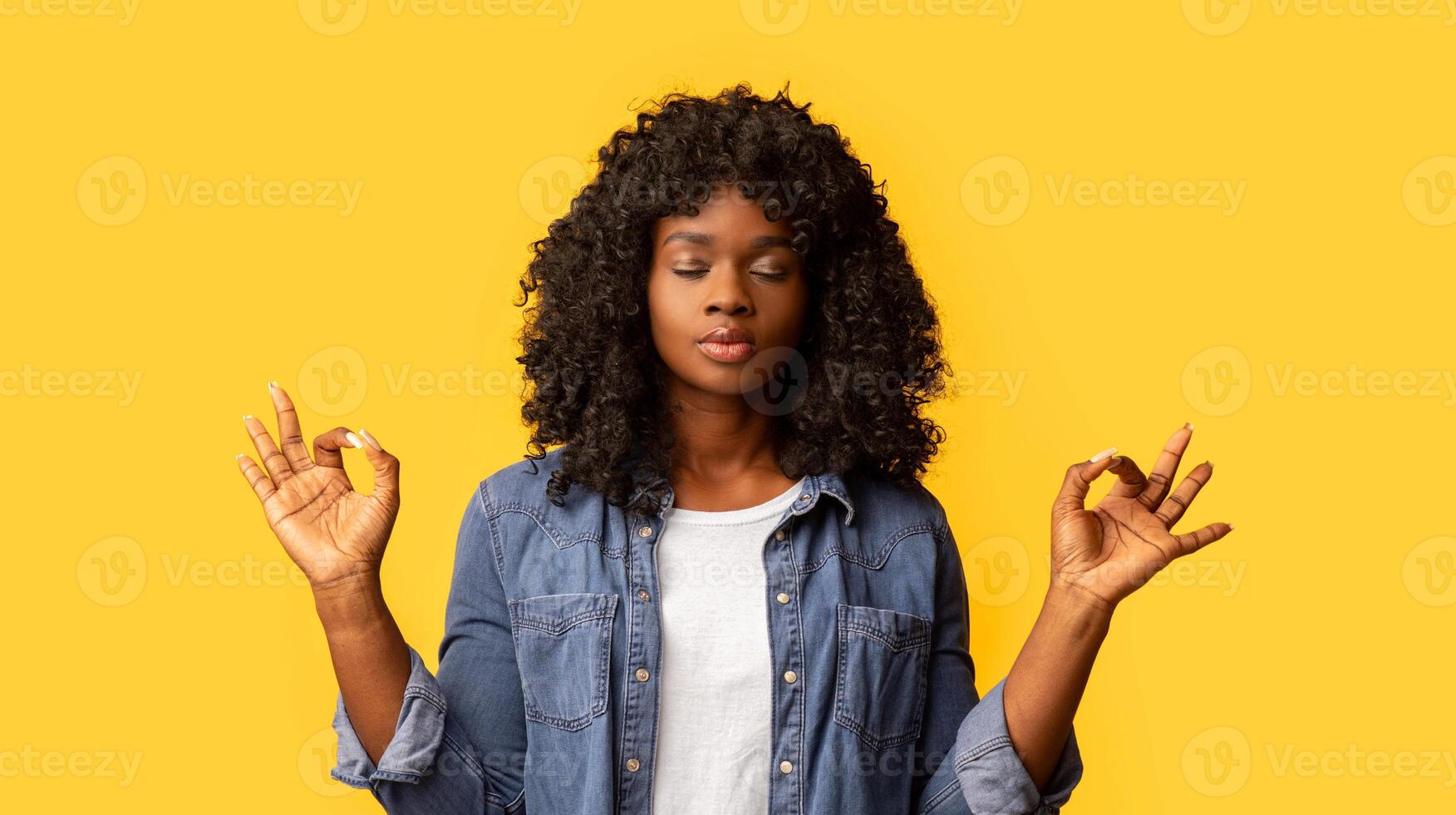 Staying calm and relaxed in urban atmosphere. Cute determined dark-skinned woman in casual outfit, closing eyes and raising hands in zen gesture, meditating or practicing yoga over yellow background photo