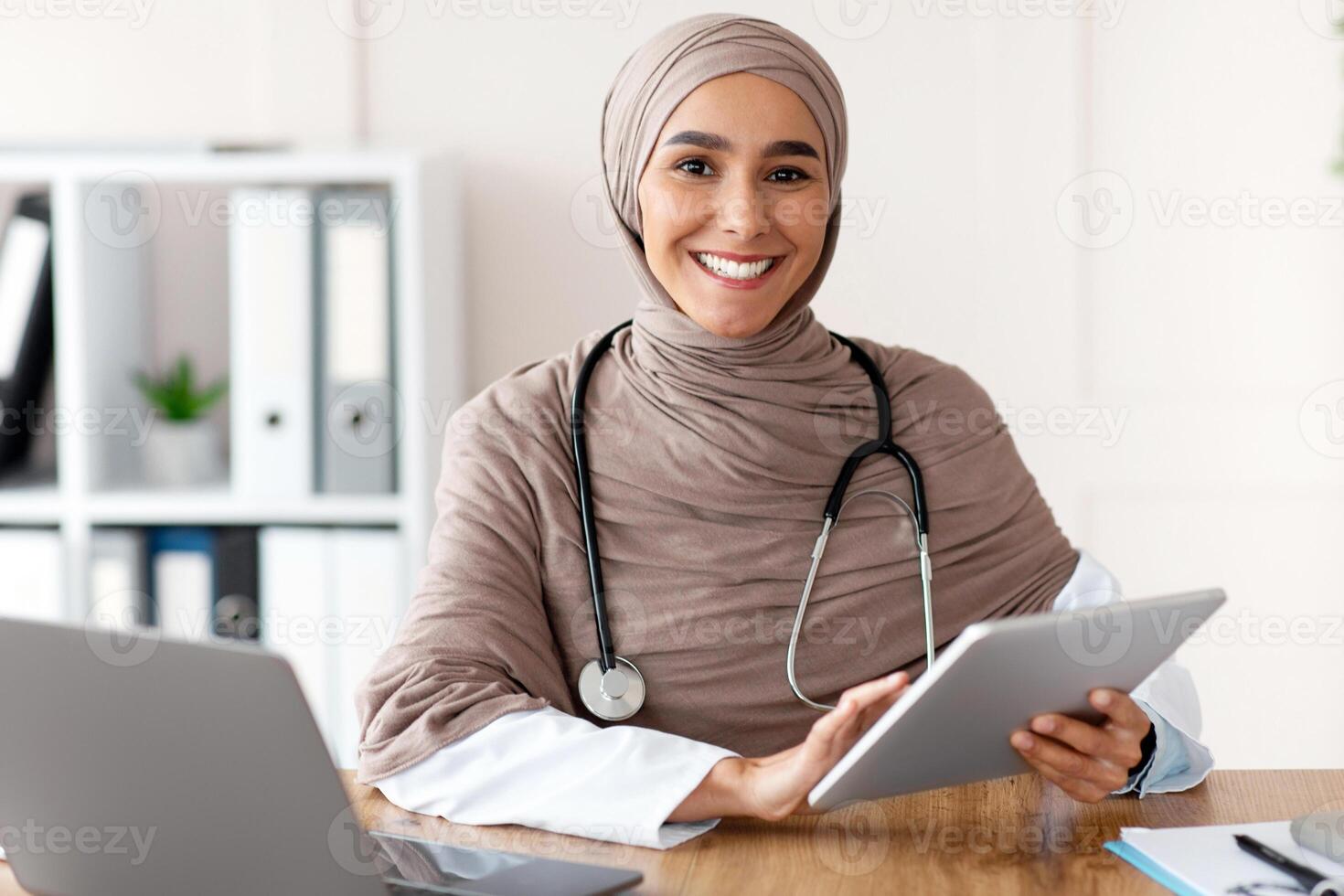 Online appointment with doctor concept. Attractive muslim woman doctor working in clinic, sitting at workdesk, using digital tablet and laptop, making online appointment with patient or training photo