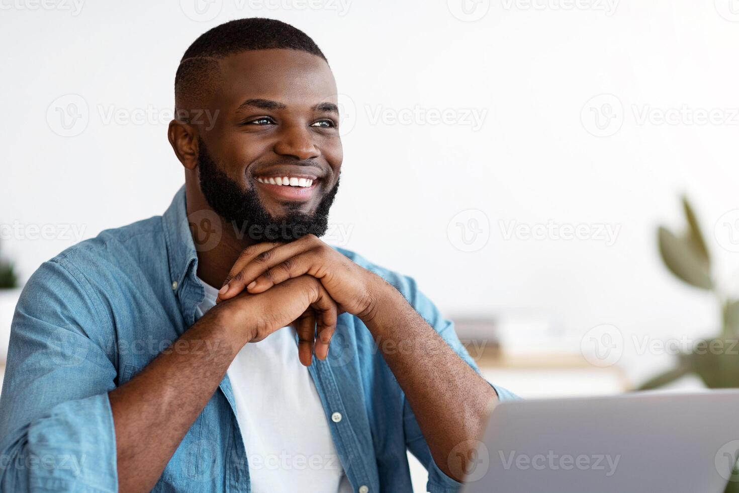 Portrait of pensive smiling black freelancer sitting at desk in home office, thinking about business strategy while working on laptop computer, looking away with pleased face expression, free space photo