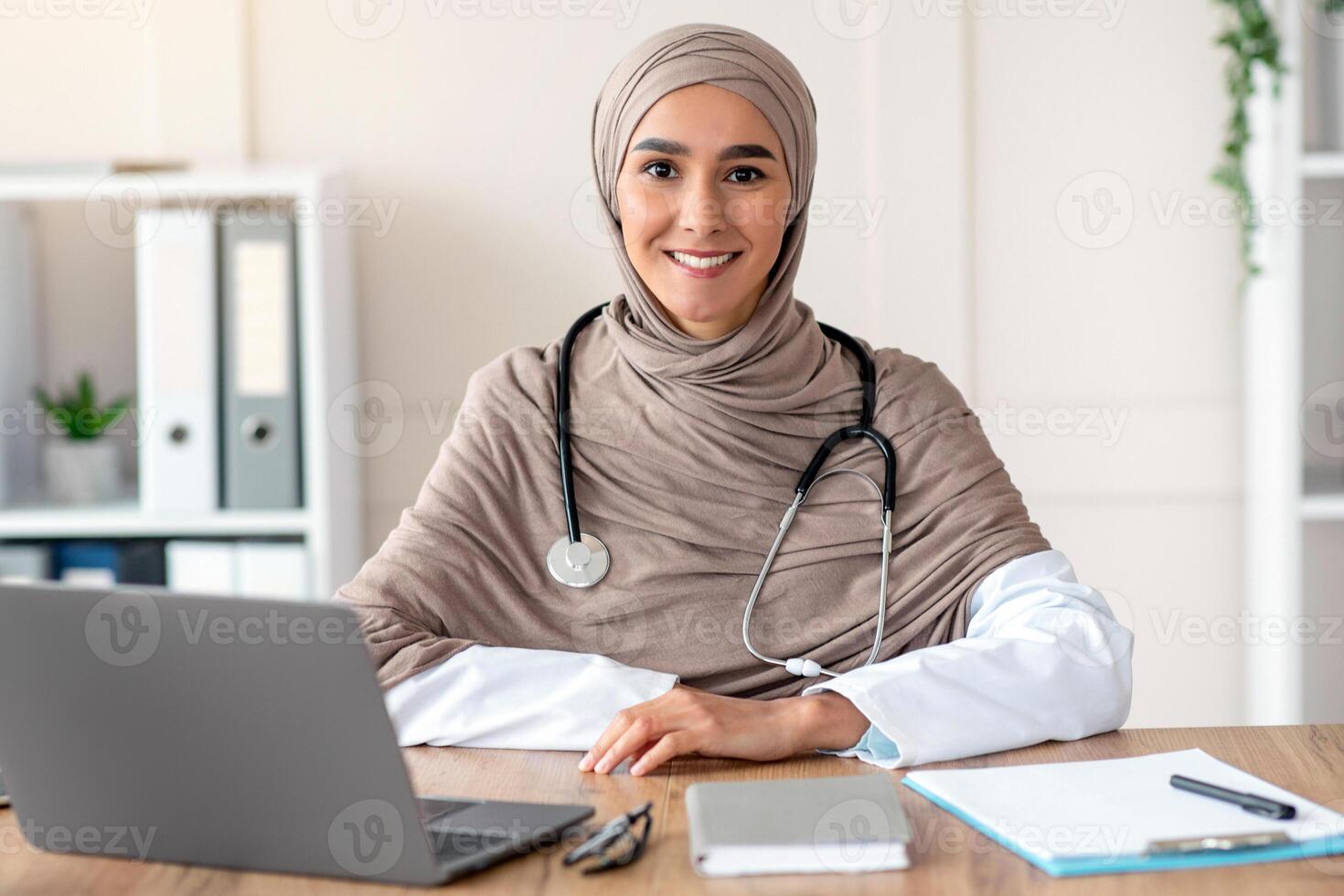 Portrait of smiling female doctor young beautiful woman in hijab with stethoscope posing in her cabinet, sitting at workdesk with medical chart, notebook and laptop in clinic, copy space photo