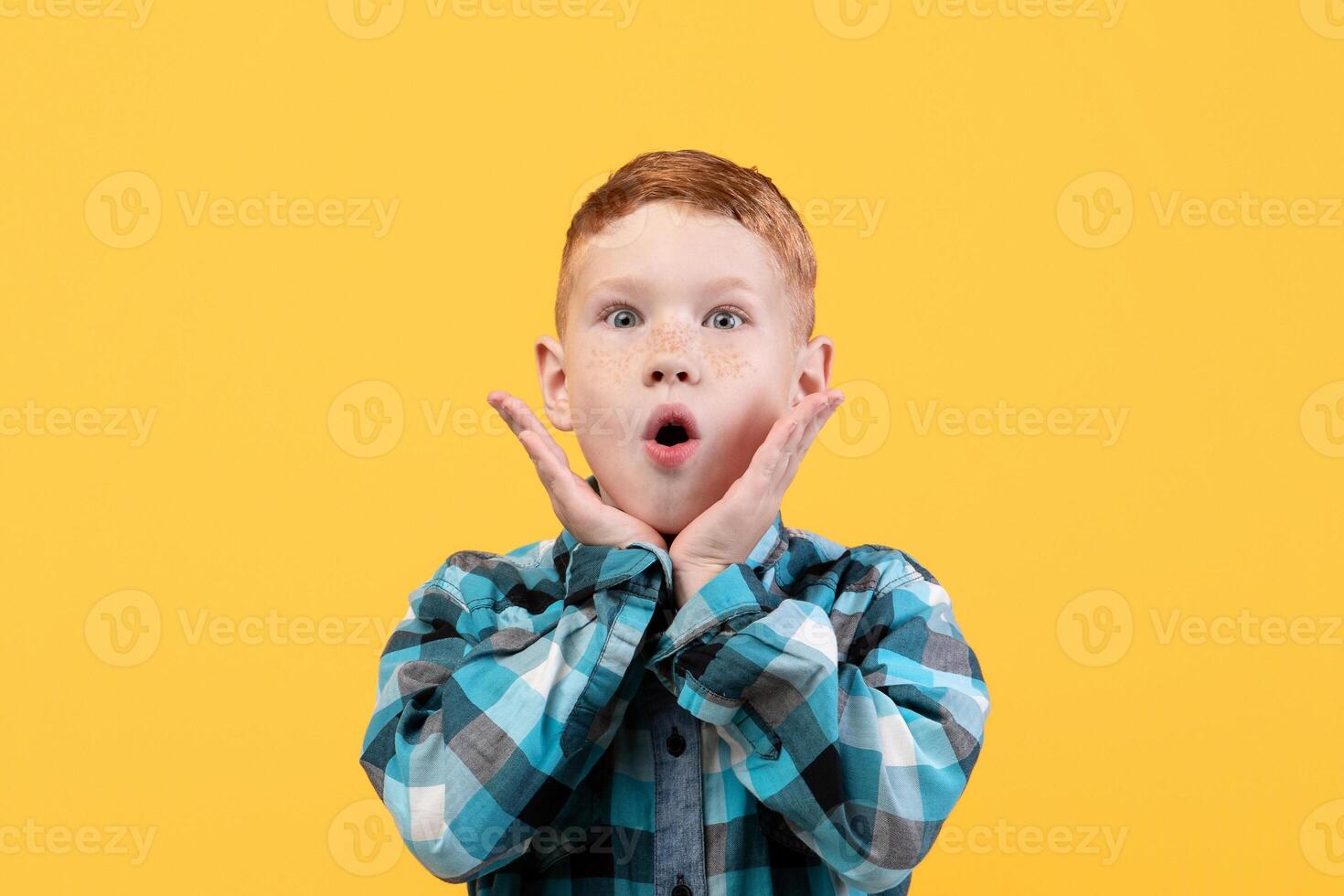 Surprised little boy in casual shirt, face expression concept. Shocked redhead kid keeping mouth open and touching his face, seeing something shocking, yellow studio background, copy space. photo